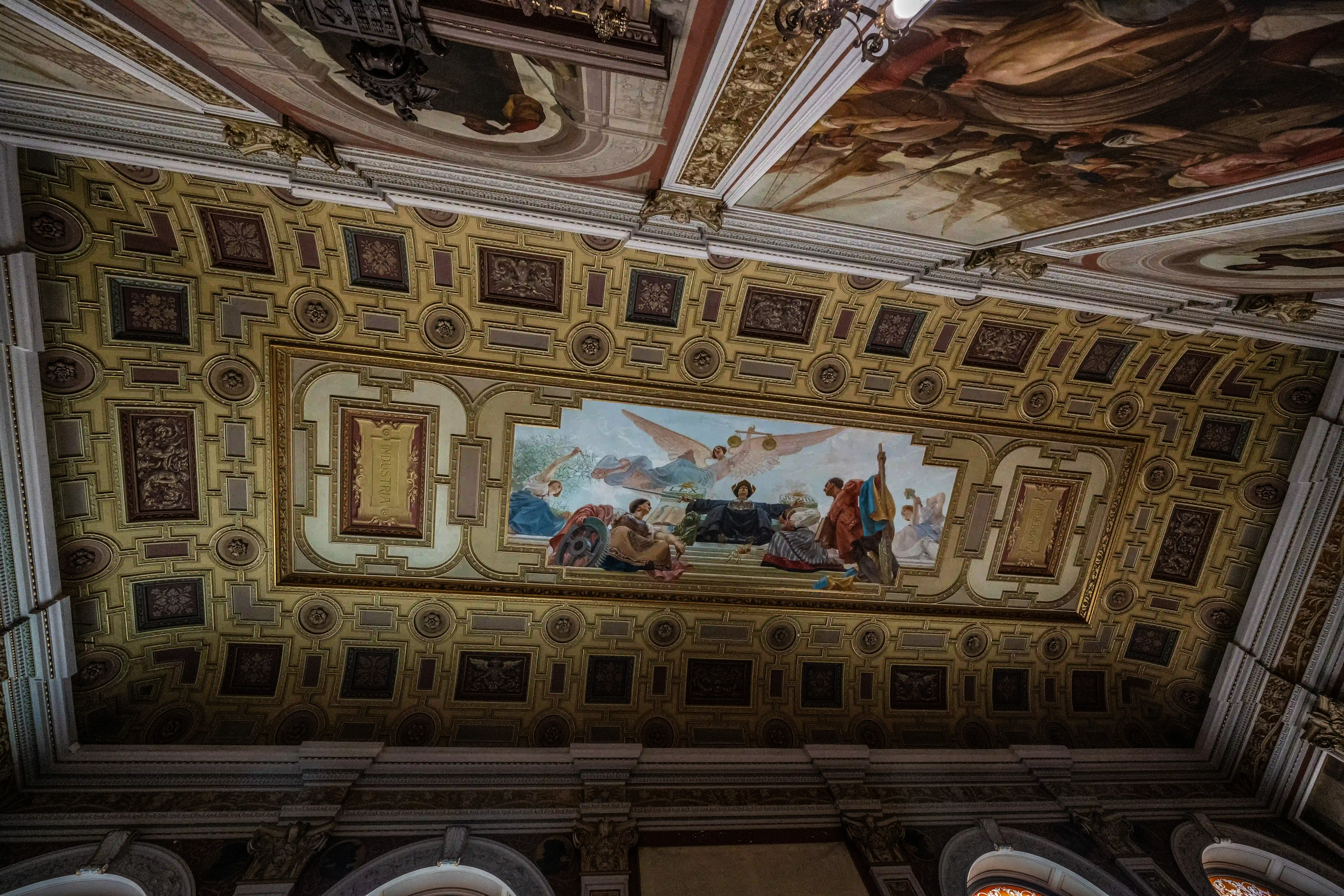 Low-angle view of a highly ornate gold ceiling with a central fresco depicting figures and an angel, surrounded by decorative patterned panels and elaborate cornices.