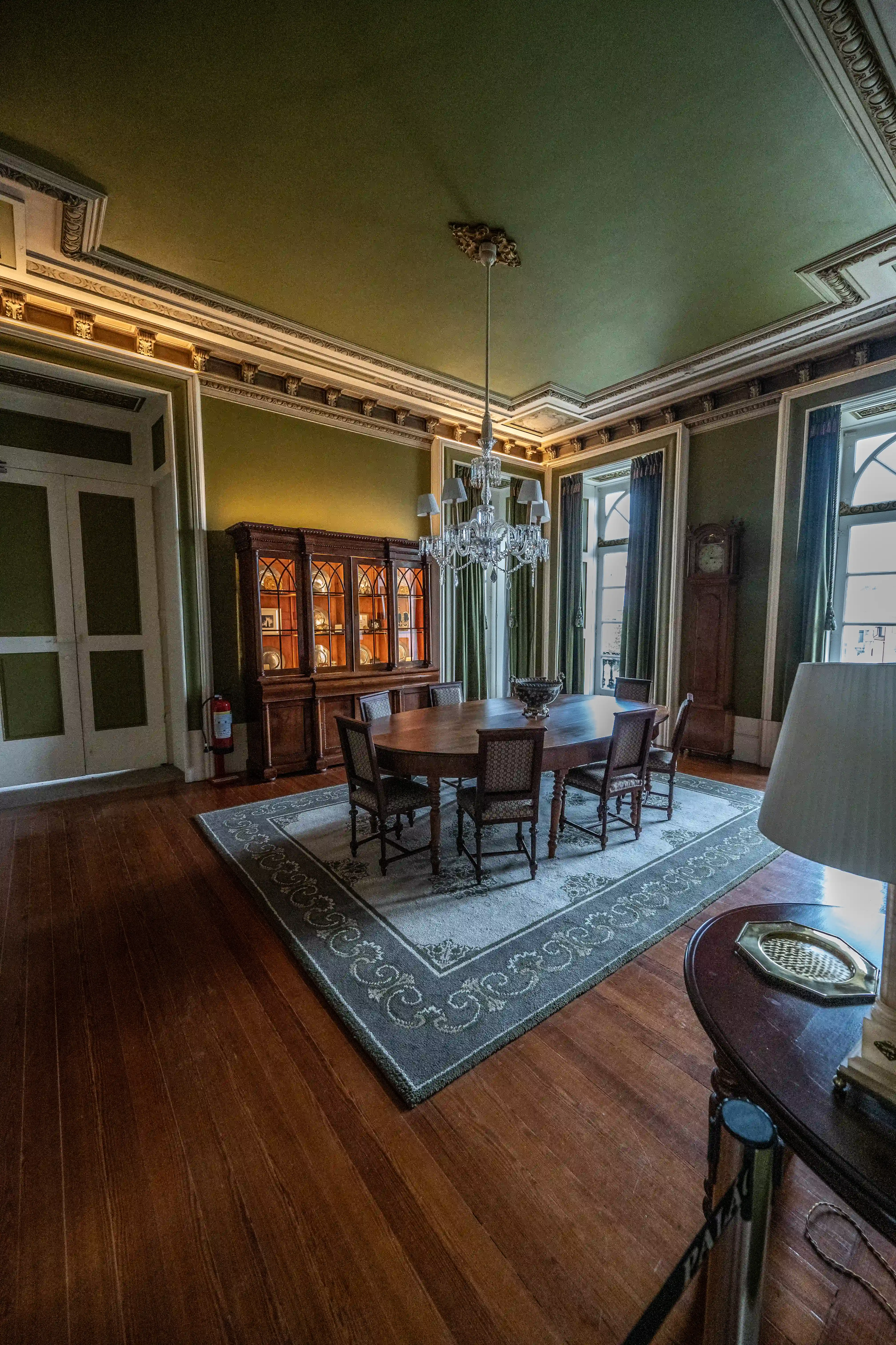 A wide-angle shot of a formal dining room featuring a large wooden table with six chairs, a crystal chandelier, a display cabinet, a grandfather clock, and a patterned rug on a wooden floor.