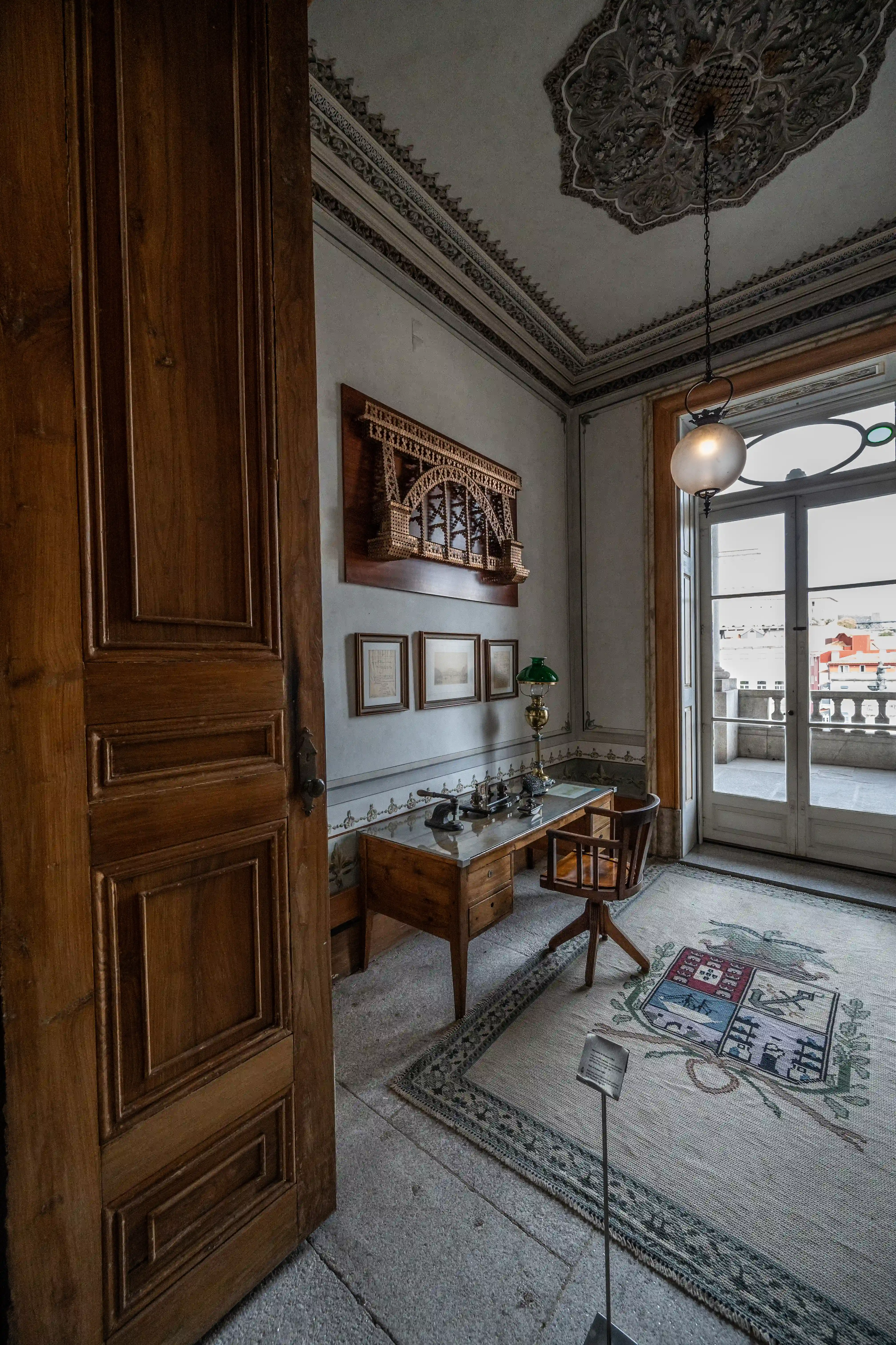 An ornate study room with a wooden desk, chair, and an antique lamp. A wooden model of a bridge is mounted on the wall above three framed pictures. A large glass door leads to a balcony with a city view. A patterned rug with a crest covers the floor.