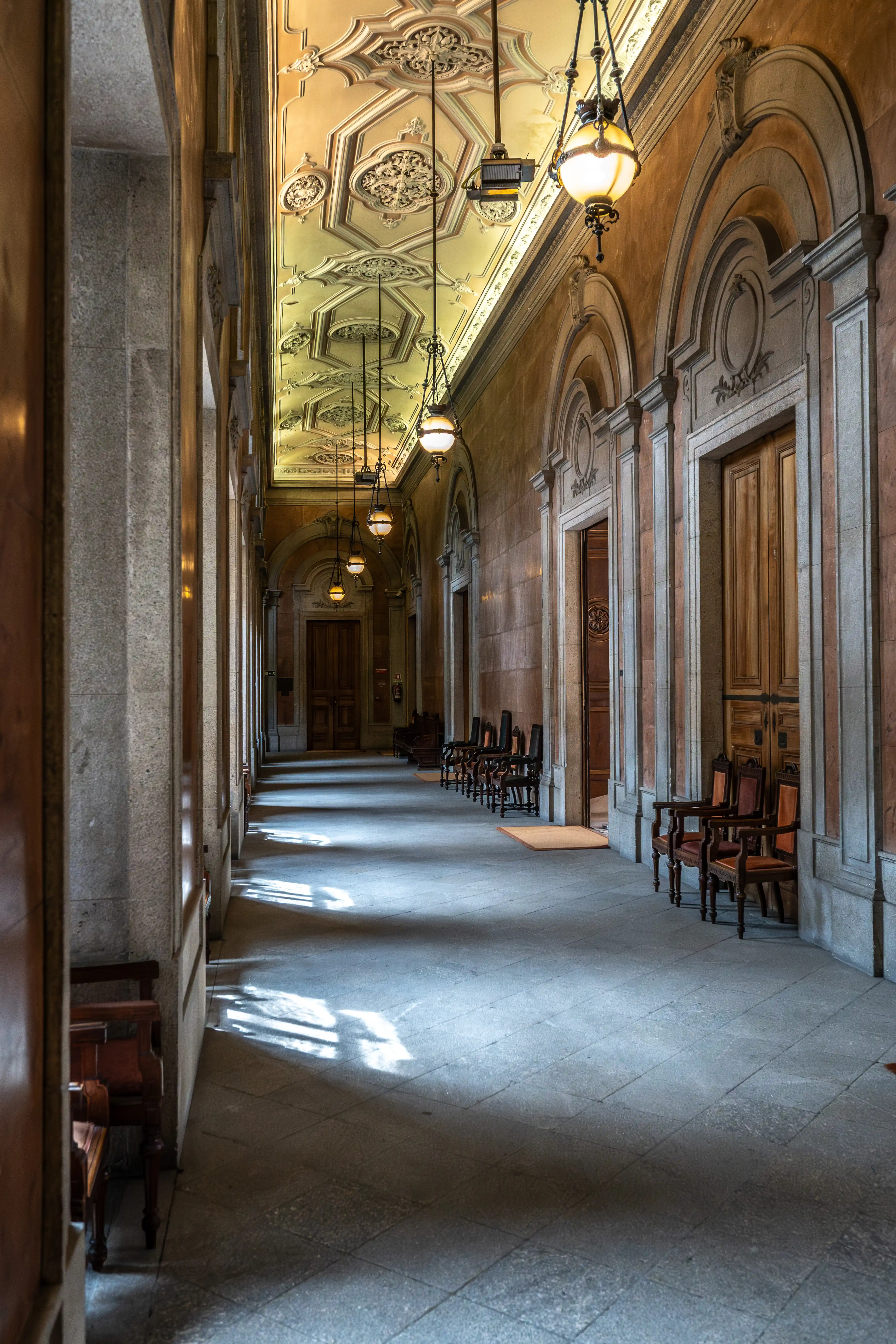A long, dimly lit hallway with a highly decorative ceiling featuring intricate mouldings and ornate hanging lamps. Sunlight casts a pattern of light and shadow across the stone floor, and dark wooden chairs line the walls.