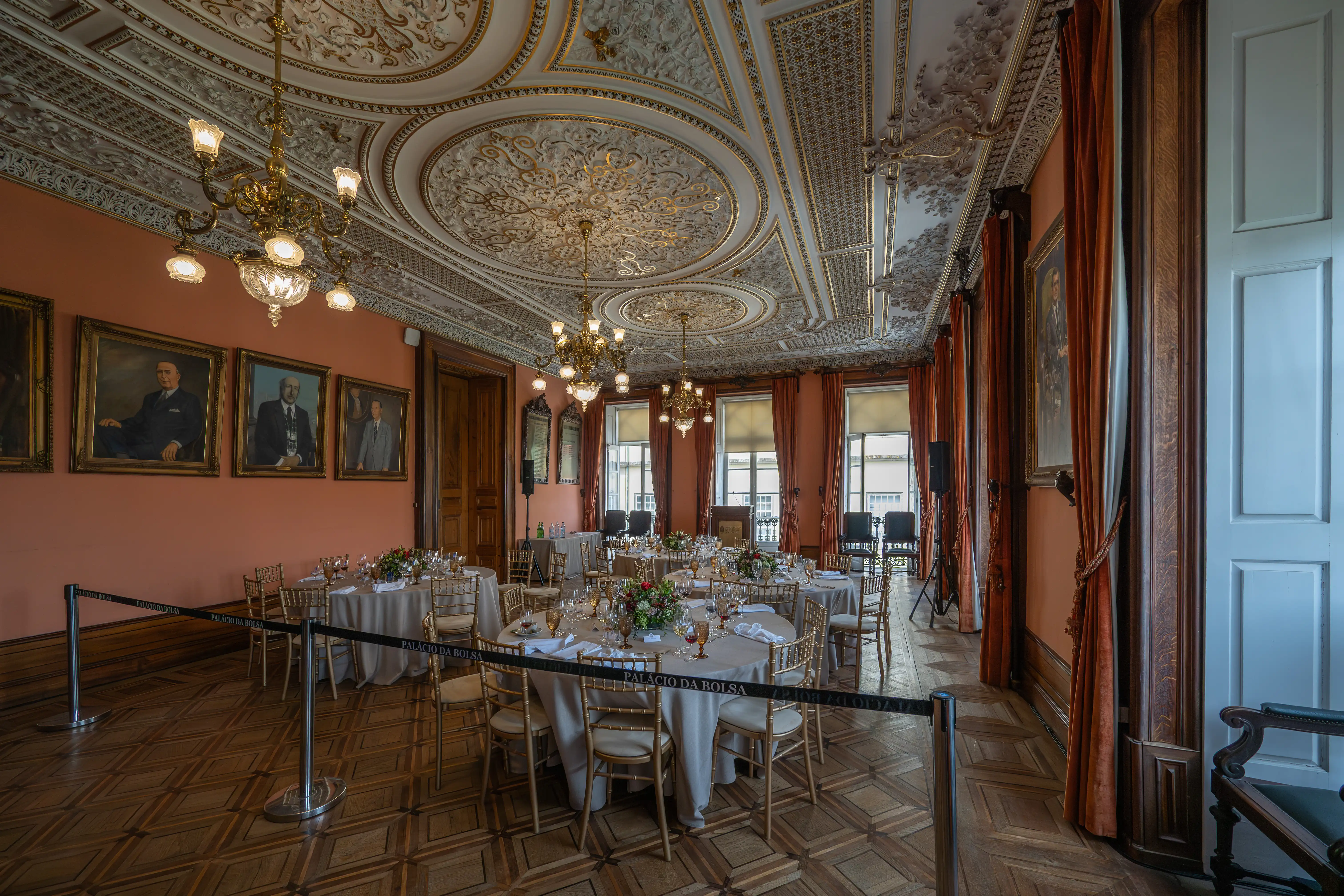 A grand dining hall in a historic building, featuring multiple round tables set for a formal dinner with floral centrepieces and golden chairs. The room is decorated with ornate gilded ceiling plasterwork, large chandeliers, and portraits on salmon-pink walls, with rich orange curtains and a polished wooden floor. The text PALÁCIO DA BOLSA is visible on a black barrier in the foreground.