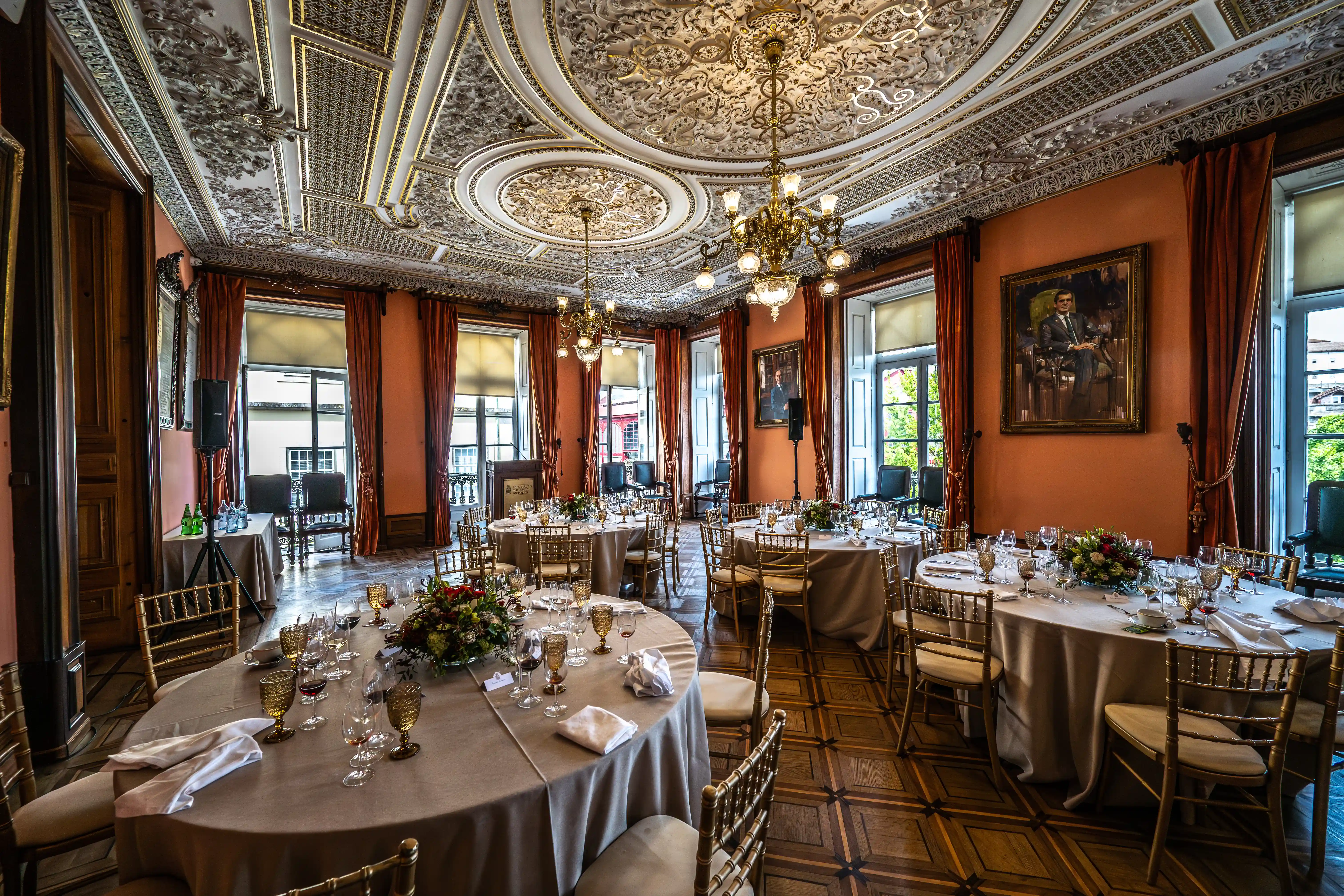 A formal dining room is set with round tables, elegant glassware, and white napkins. The room features an ornate gold-detailed ceiling with chandeliers, burnt-orange curtains, a portrait on the wall, and a decorative wooden floor. Gold chairs surround the dining tables.
