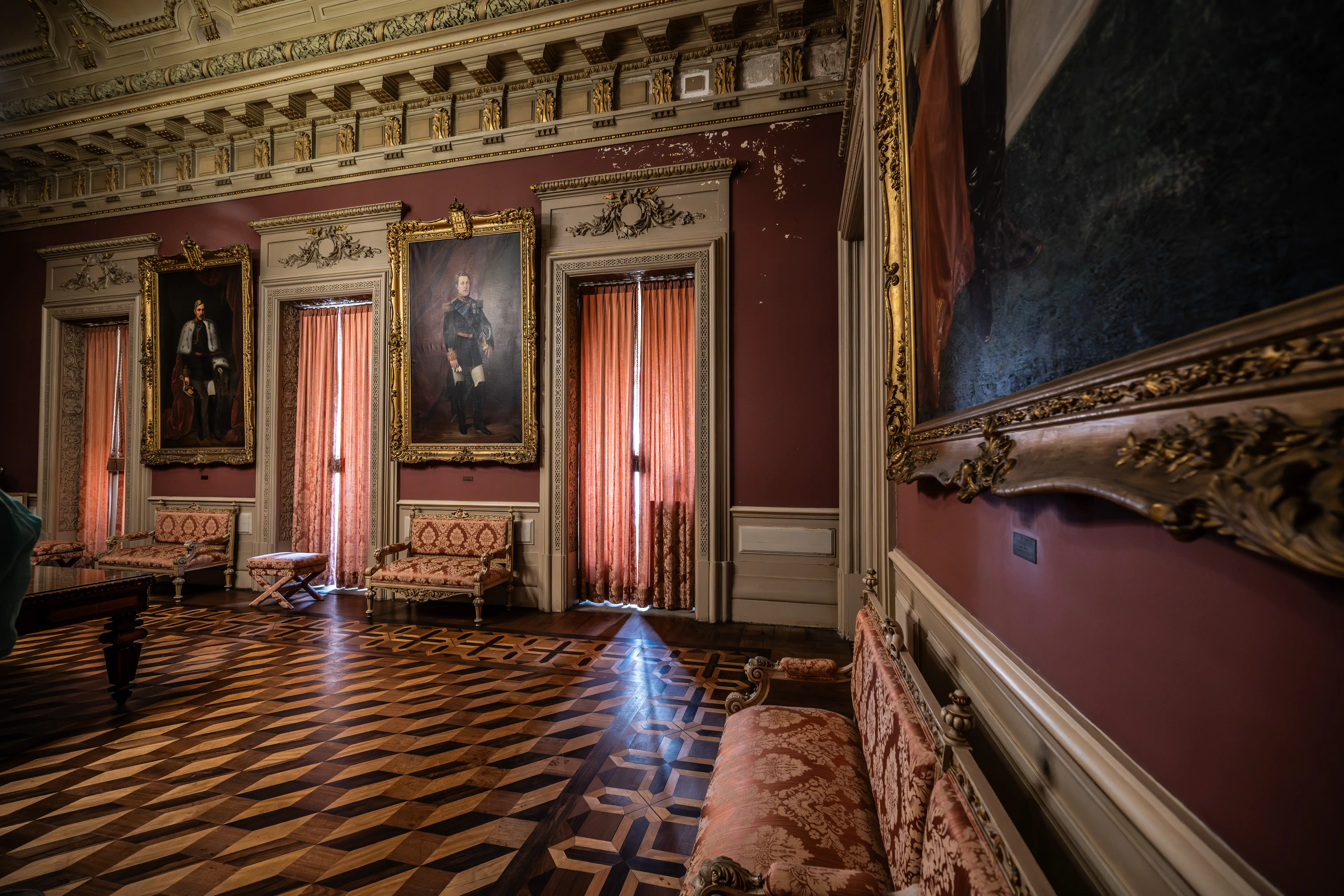 A wide shot of a richly decorated room with dark red walls, featuring several large, framed oil paintings and ornate, gilded furniture. The floor is made of dark and light wood arranged in a striking geometric parquet pattern, with a beam of light shining across it from behind coral-coloured curtains.