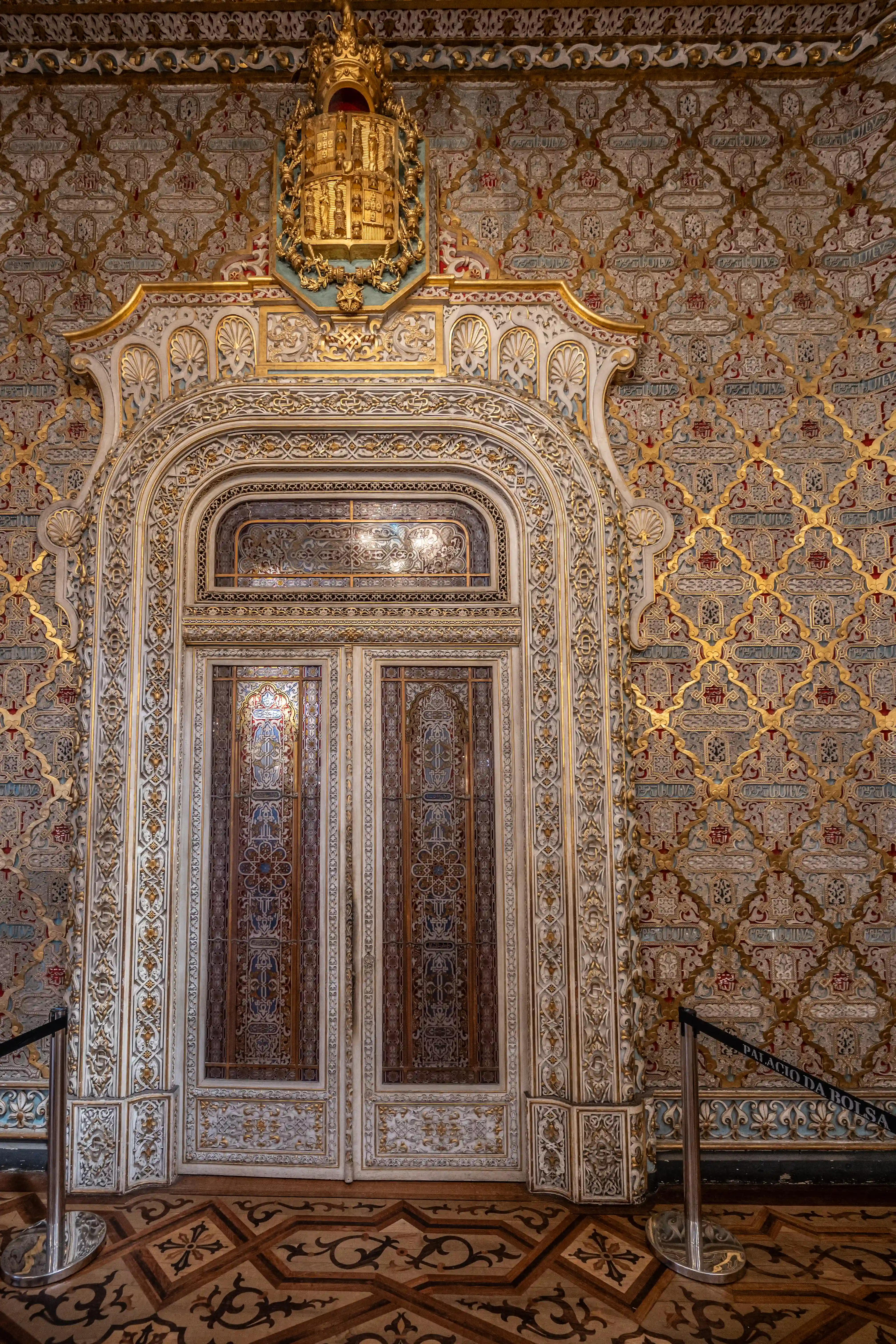 A highly ornate double door with decorative glass panels and intricate gold detailing. Above the door, a golden royal crest is mounted on a turquoise background, all set against a wall covered in a detailed gold and blue Arabesque pattern. The floor is a mosaic of patterned wood.