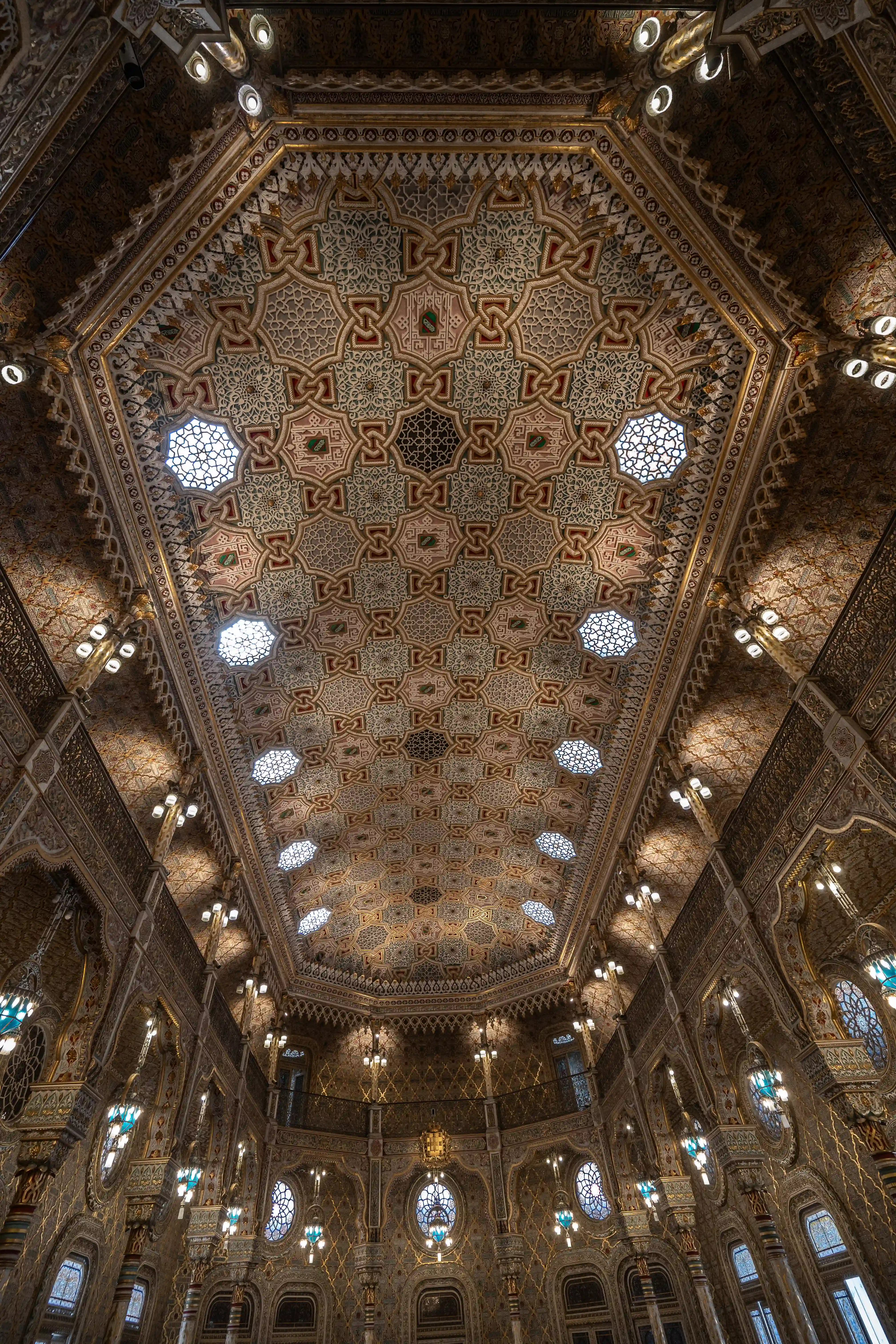 A low-angle view of a highly decorated, octagonal ceiling adorned with intricate geometric patterns in gold, red, and white, interspersed with small, lattice-covered skylights. Ornate lamps hang from the elaborately patterned walls below.
