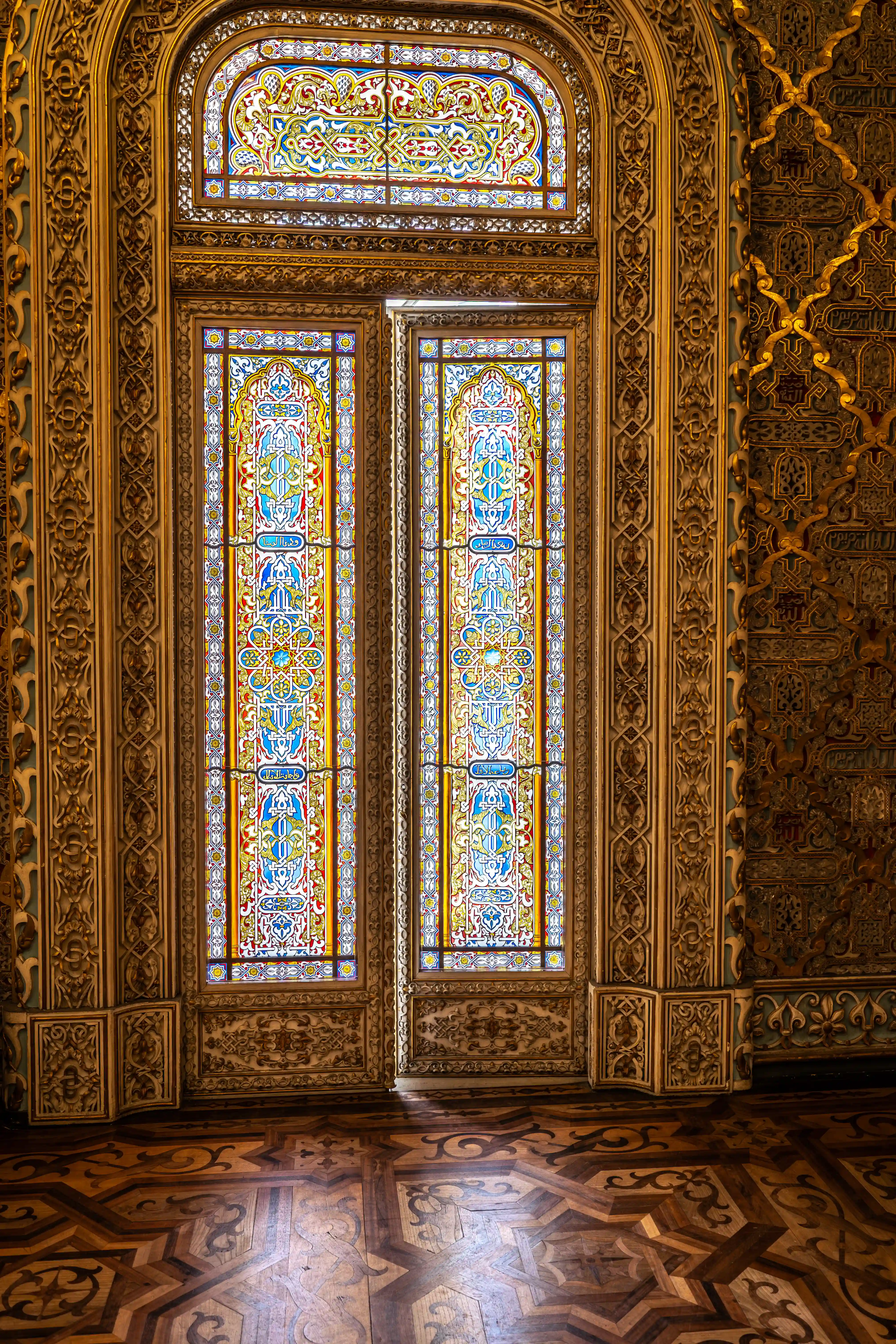 A close-up view of tall, double doors with arched stained-glass panels in vibrant blues, yellows, and reds, framed by ornate golden carvings. Above, a similar arched stained-glass window. To the right, a wall features intricate golden Moorish patterns and calligraphy. The foreground shows a detailed, geometric wooden parquet floor.