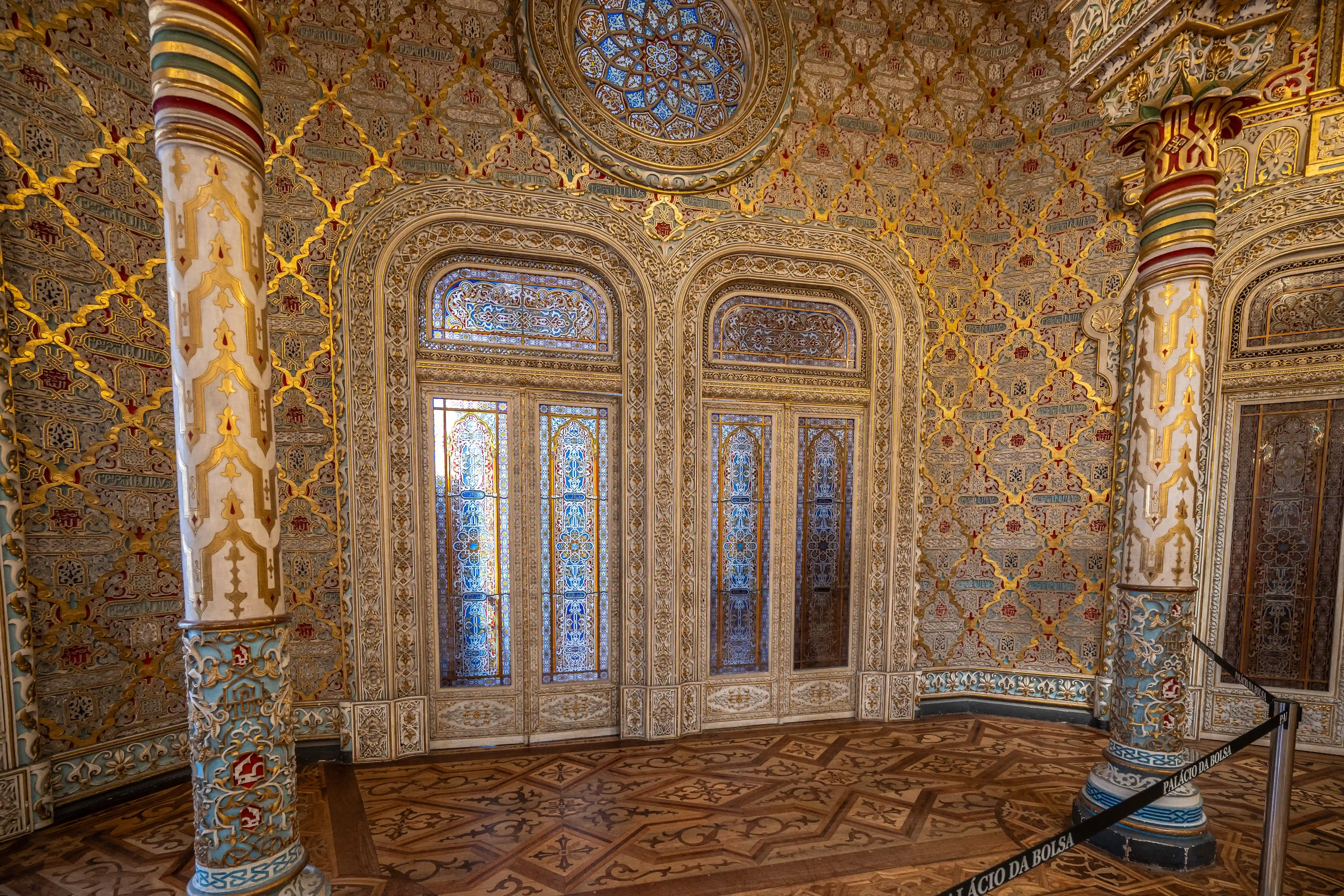  Interior of a richly decorated room at the Palácio da Bolsa, featuring walls with elaborate gold and colourful Mudejar patterns, two large arched doorways with stained glass, and ornate columns. The floor has a detailed wooden parquet.