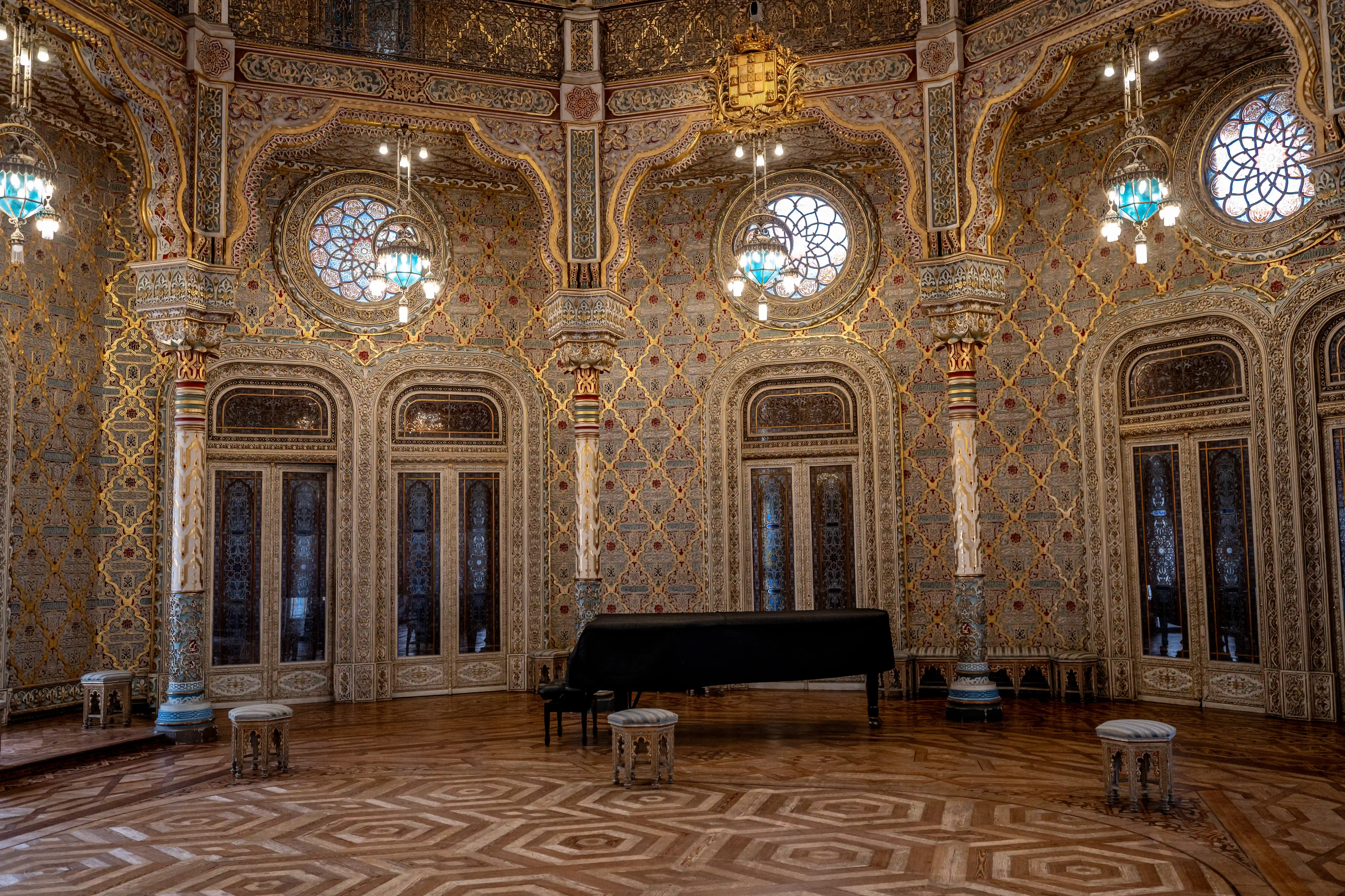 A grand hall with walls covered in intricate gold and red Mudejar patterns, featuring arched doors, circular stained-glass windows, and ornate golden trim. A black-covered grand piano sits centrally, with small decorative stools and a patterned wooden floor visible.