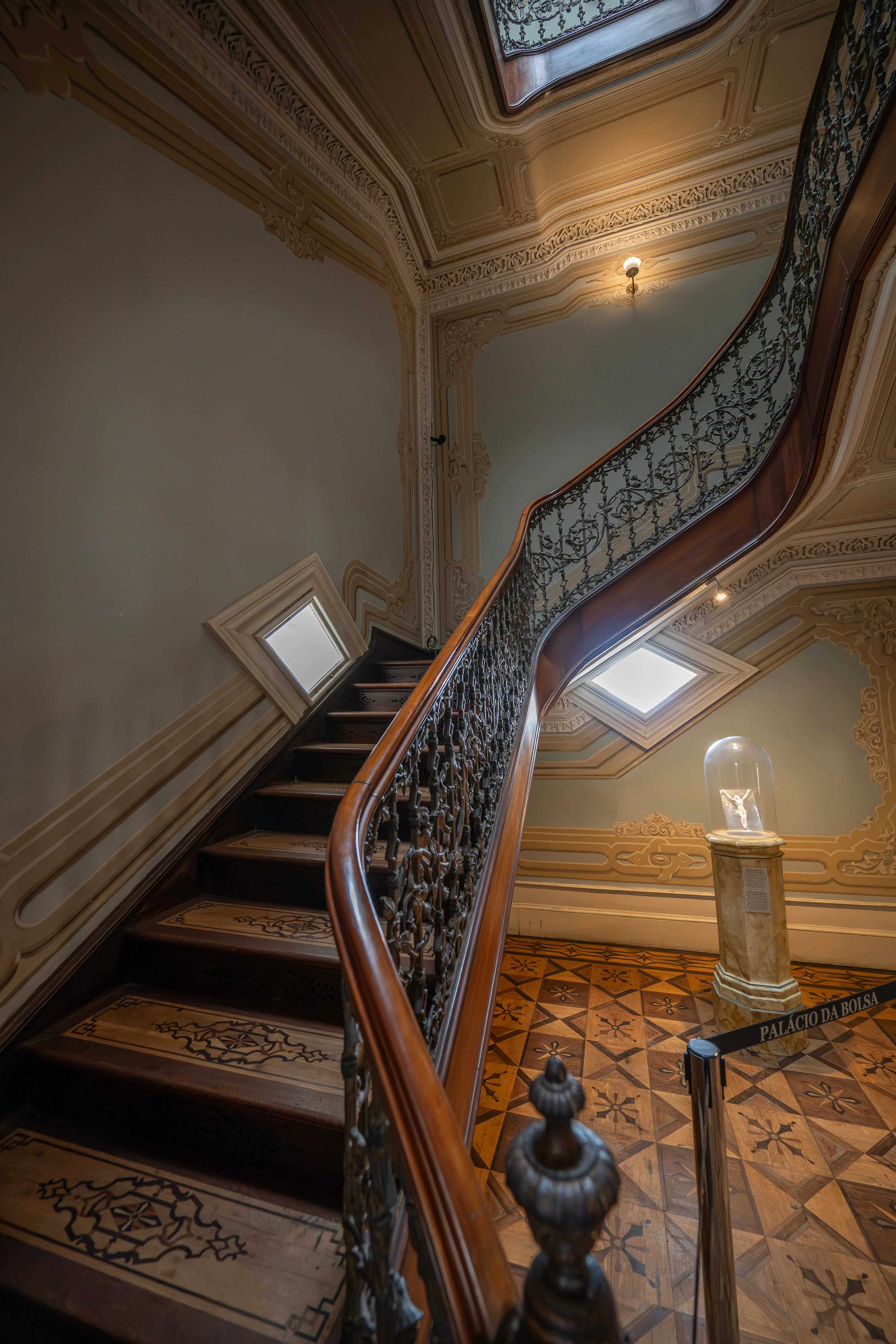 A detailed photograph of an opulent, curved wooden staircase with a decorative wrought-iron balustrade. Intricate wooden inlays adorn the steps, and elaborate plaster mouldings frame the walls and ceiling. Skylights provide natural illumination, and a display encased in glass rests on a marble pedestal, with a cordon reading PALÁCIO DA BOLSA.