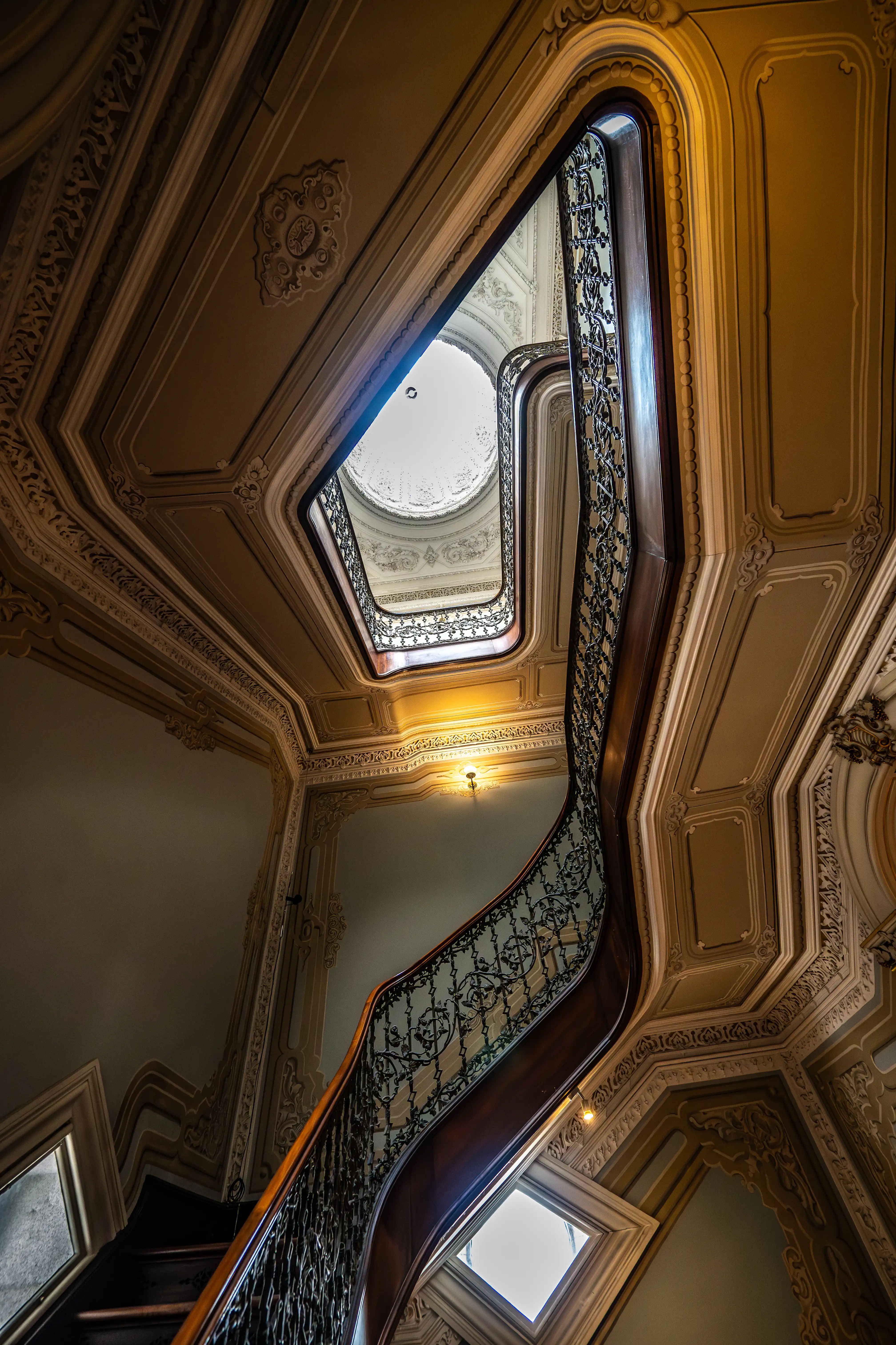 Looking up at an ornate, curved staircase with a dark wooden handrail and intricate wrought-iron balustrades, leading to a circular skylight. The walls and ceiling feature detailed decorative plaster mouldings.
