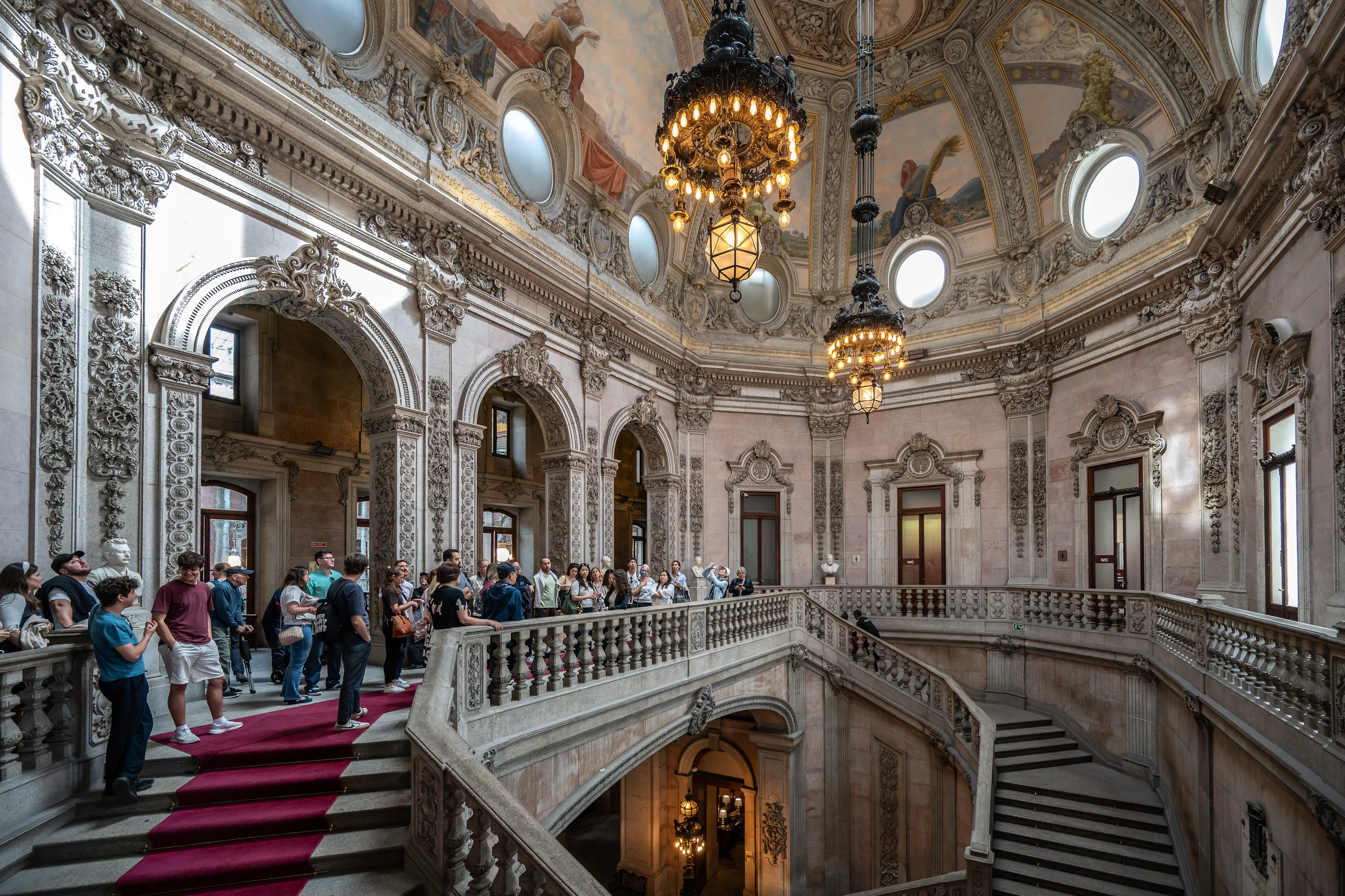 A wide shot of a grand indoor staircase with a red carpet, ornate stone carvings, and large decorative chandeliers hanging from a painted ceiling. A group of people are gathered on the stairs and balcony, looking around.