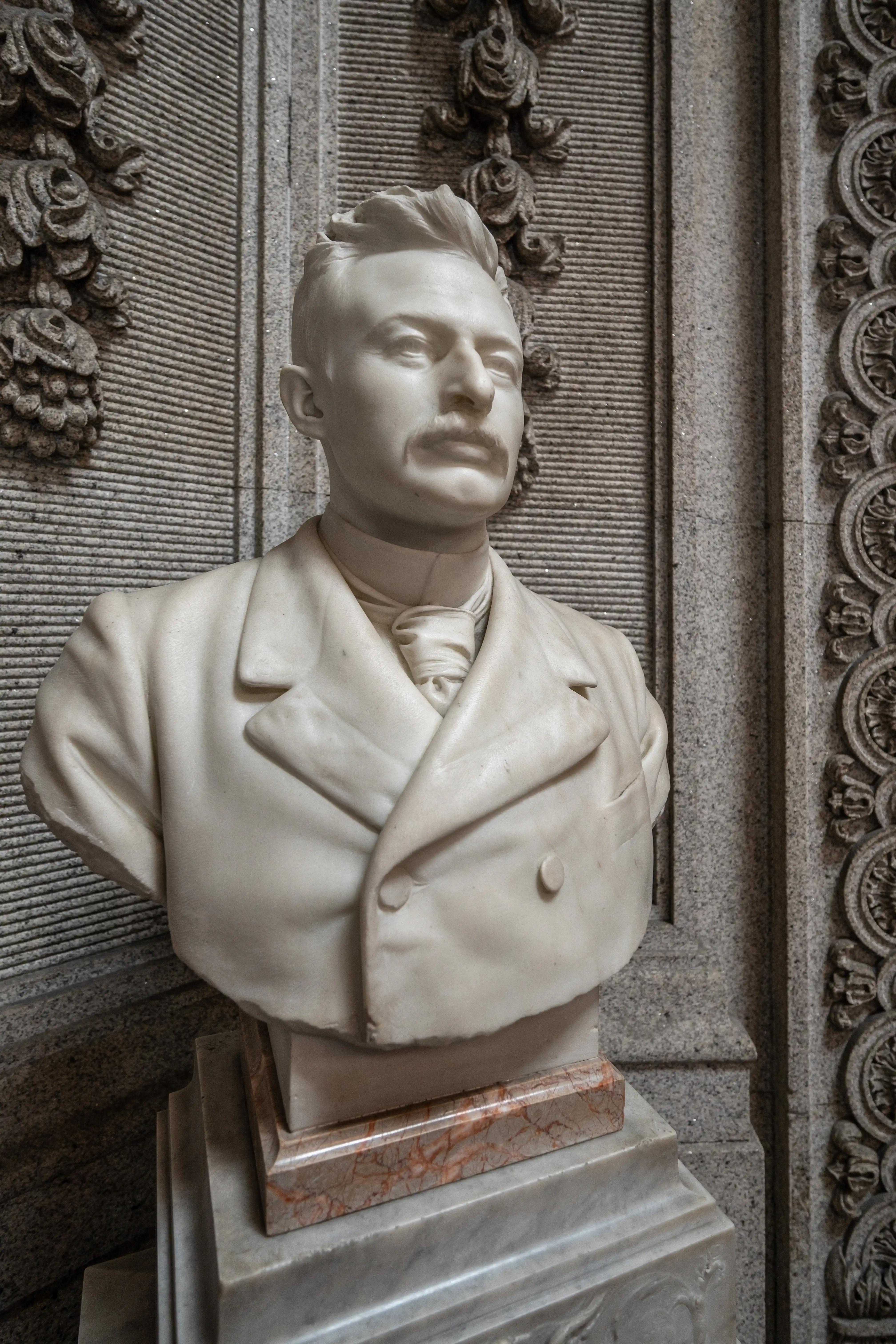 A detailed marble bust of a man with a moustache, wearing a suit and tie, set against a decorative stone background.