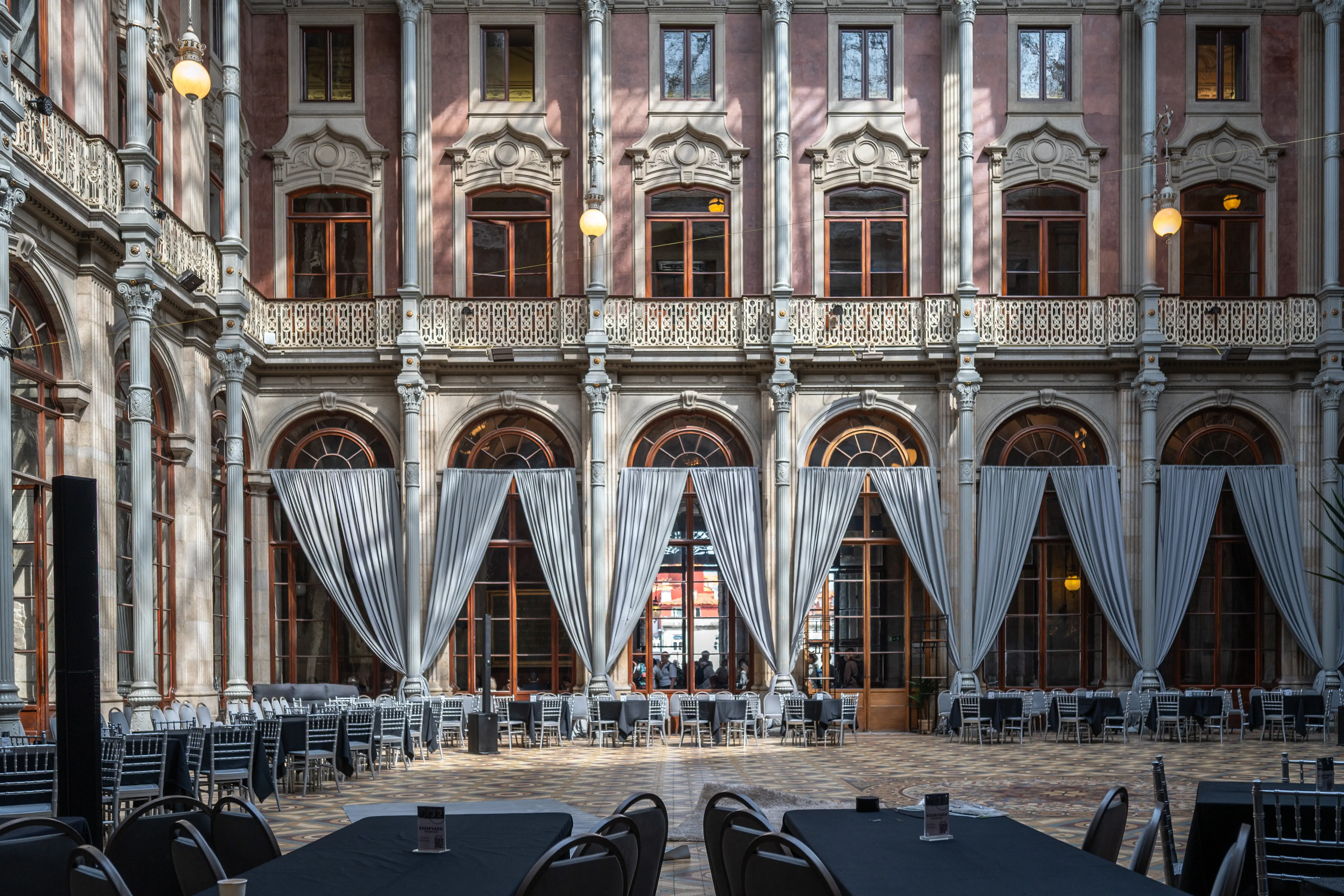 A wide shot of an ornate indoor courtyard, decorated for an event with rows of black-covered tables and chairs. Large windows with arched tops are draped with grey curtains. The architecture features multiple levels, decorative columns, and balconies with intricate metalwork.
