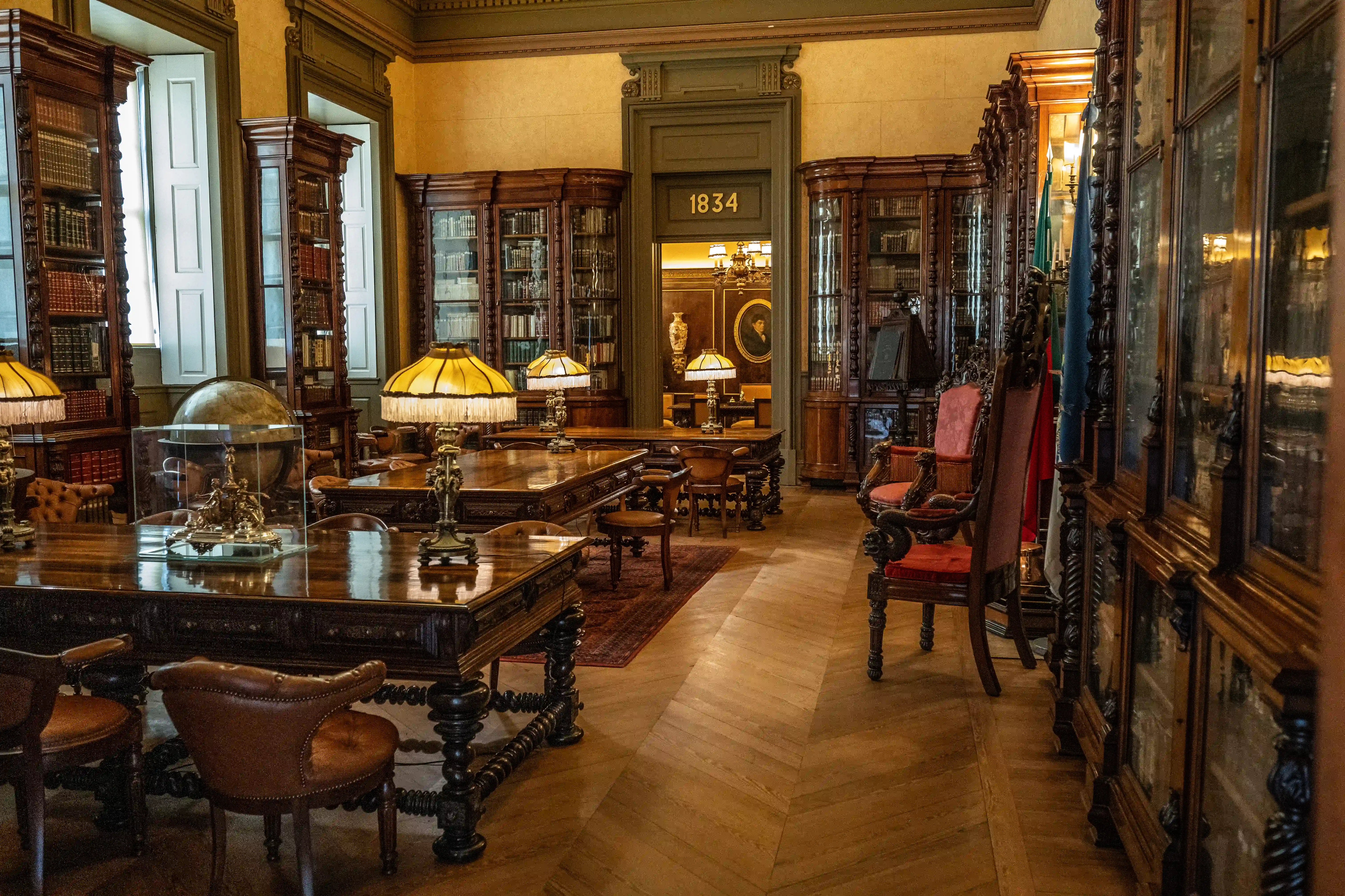 A grand library interior showcasing tall, dark wood bookcases filled with books, ornate desks with vintage lamps, and leather armchairs, with a doorway marked '1834' in the background.