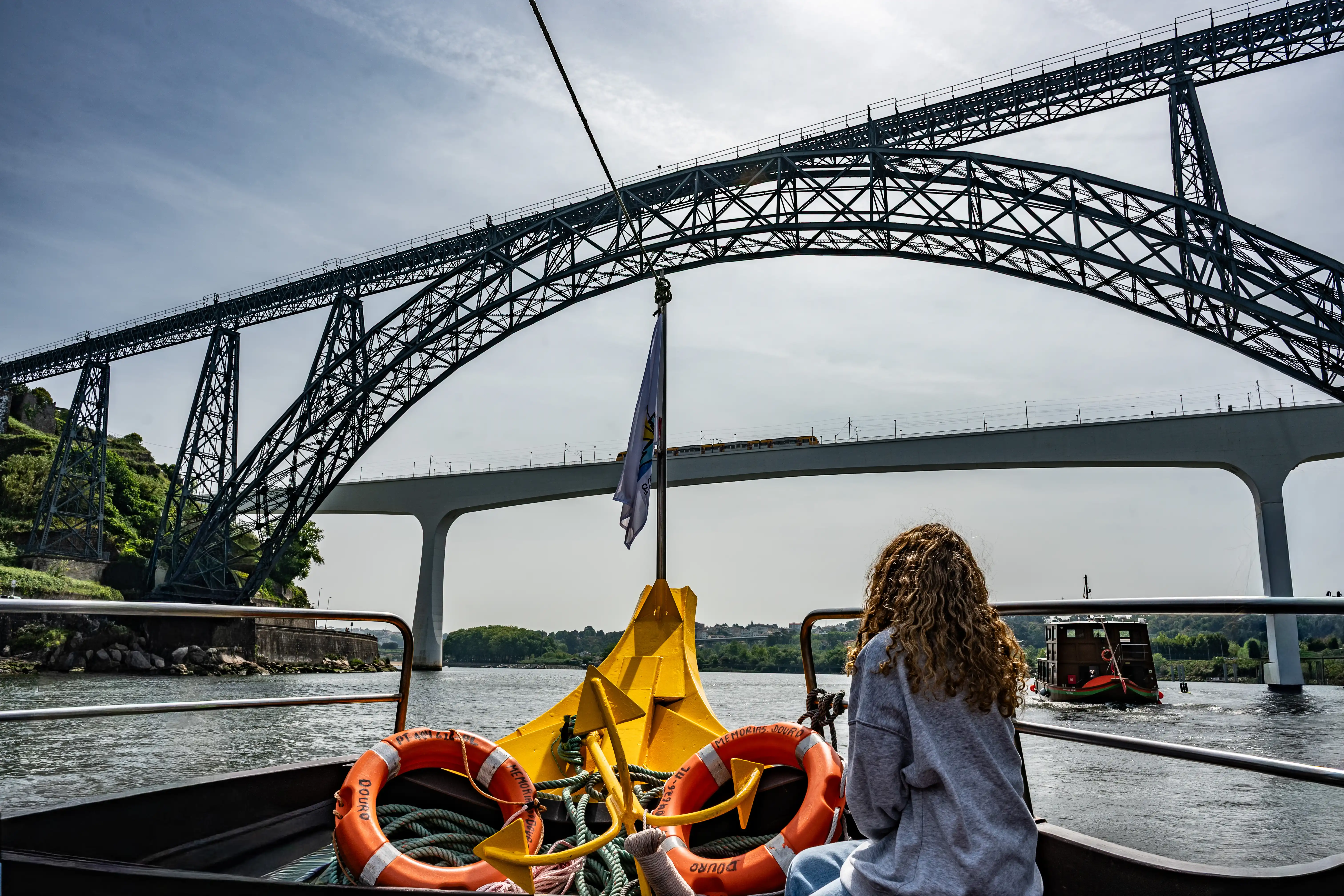 A person on a boat on the Douro River looks towards the Dom Luís I Bridge, with another boat and a train visible in the distance.