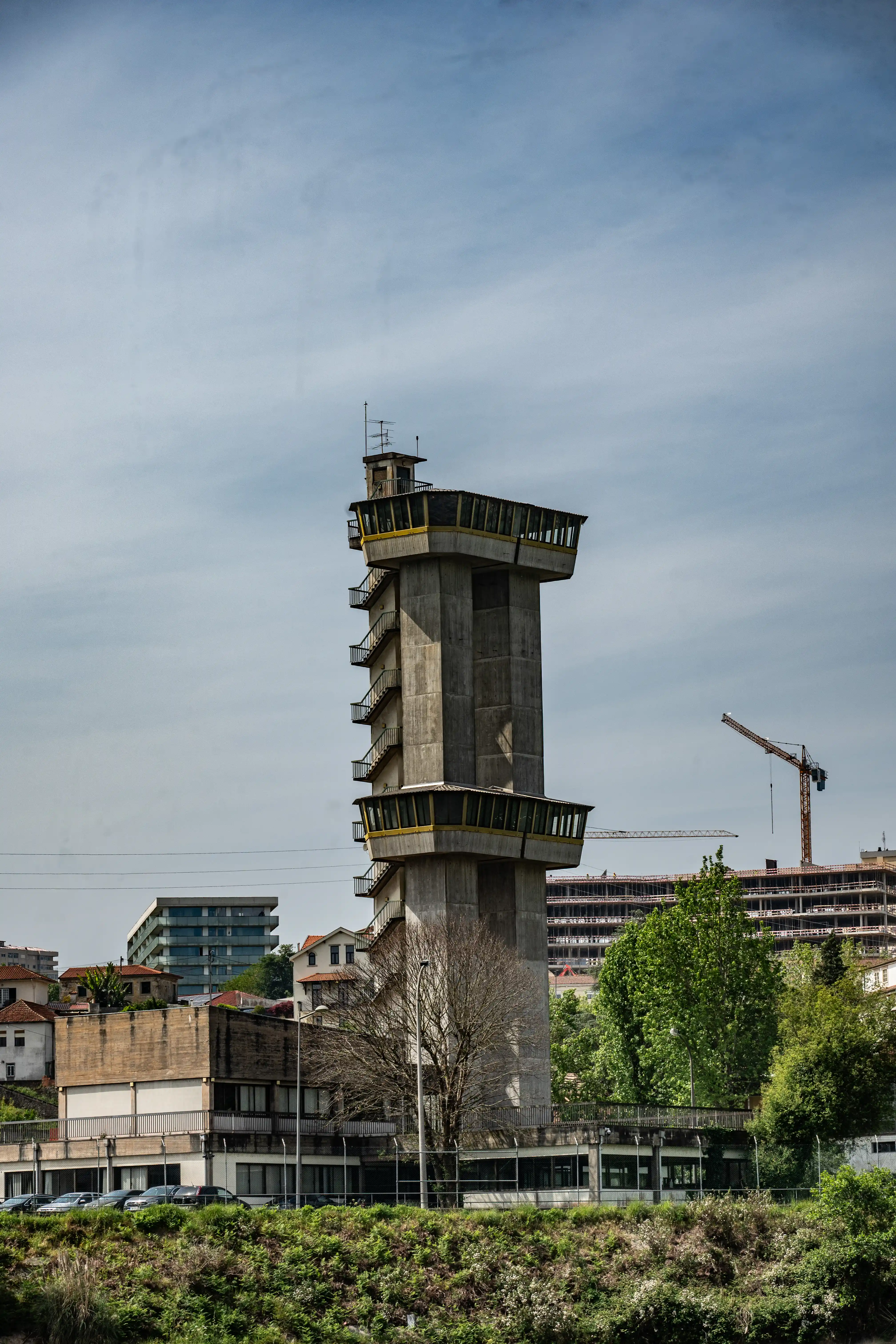 A tall, grey concrete control tower with yellow window frames and external staircases, set against a pale blue sky. In the background, several city buildings are visible, including a modern glass-fronted building and a large building under construction with a crane. The foreground is filled with green foliage and shrubs.