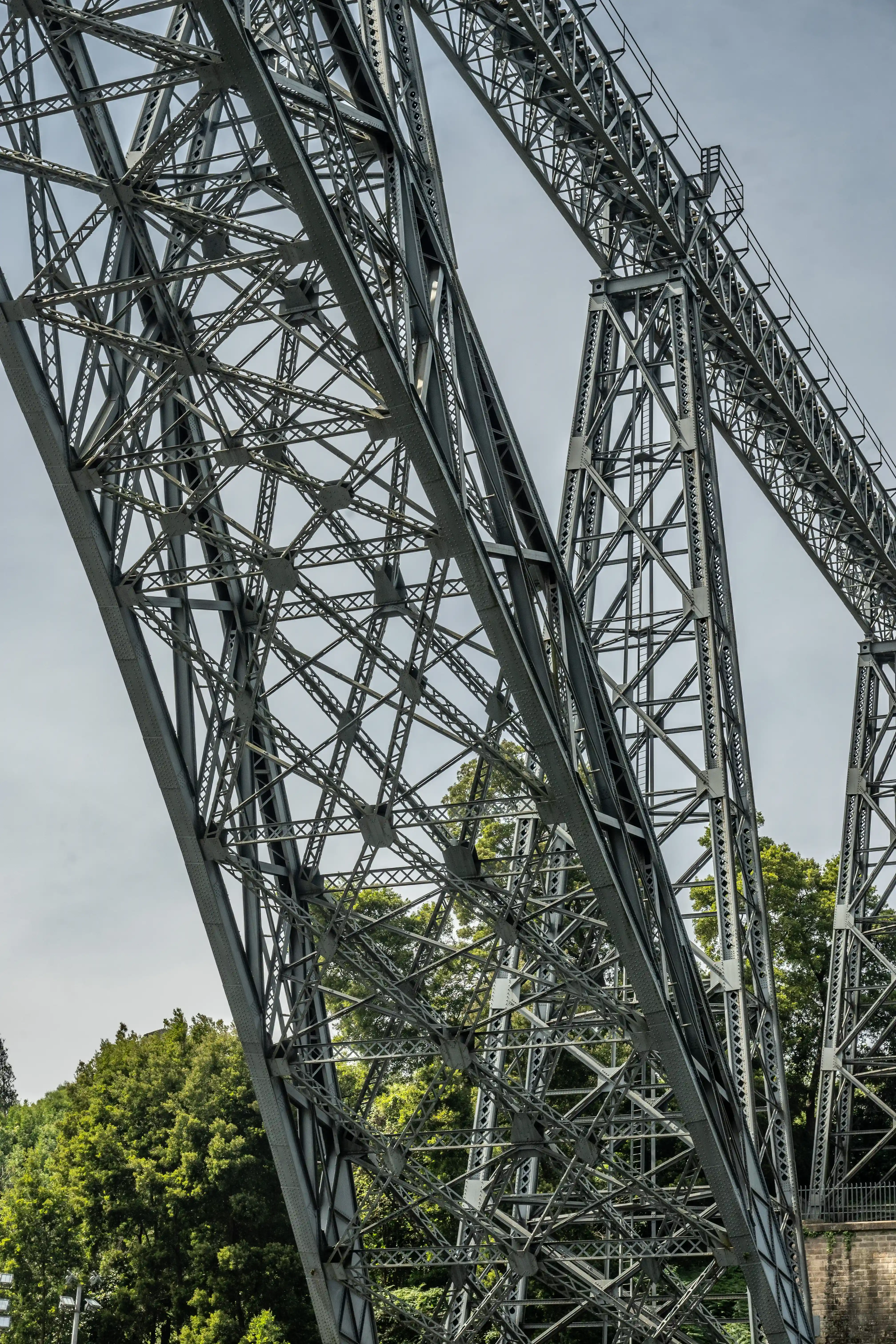 A close-up photograph from below of the complex, crisscrossing steel beams and girders of the Ponte Maria Pia. Green trees are visible on the lower left and right edges, with a grey, overcast sky overhead.