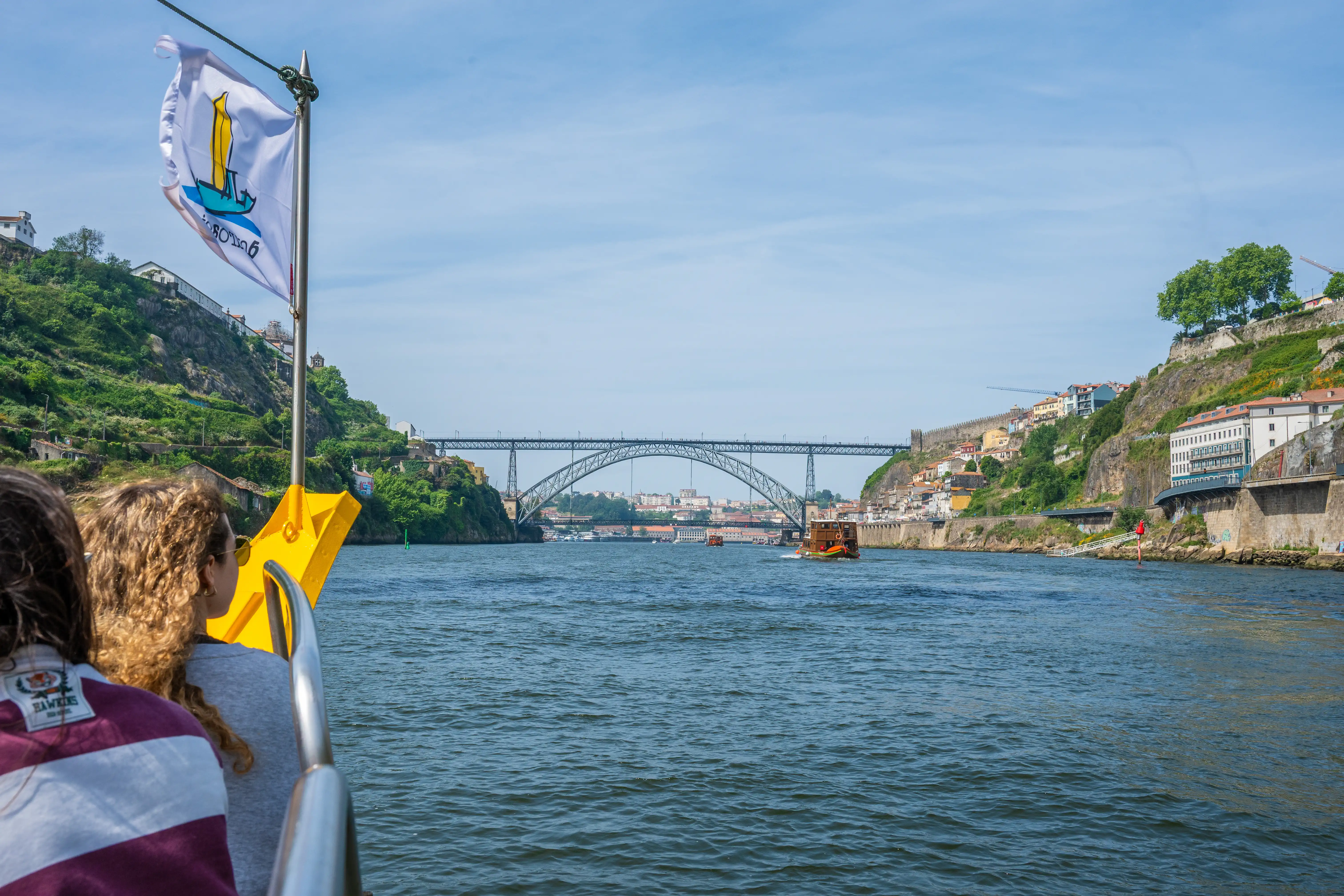 View from a boat on the Douro River, Portugal, with the Dom Luís I Bridge in the background. The image shows the hilly city landscape of Porto on the right and Vila Nova de Gaia on the left, with traditional Rabelo boats on the water. Two passengers are visible in the foreground, enjoying the scenic river cruise.