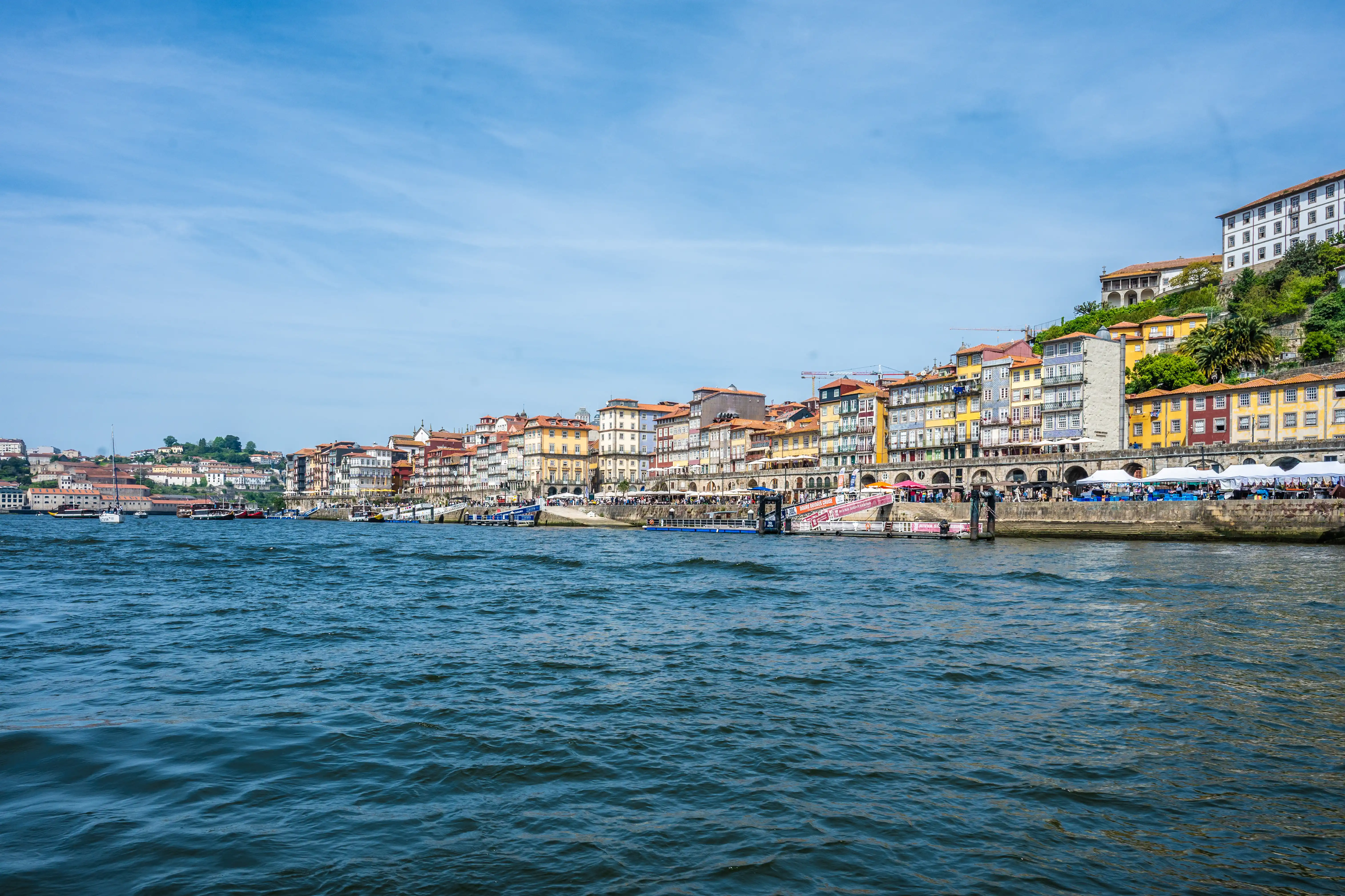 A panoramic view of the colourful buildings of Porto's Ribeira district situated along the Douro River. Boats are docked by the waterfront promenade, where people are seen dining under tents and near cafes. The sky is blue with light clouds, and the river has gentle waves.