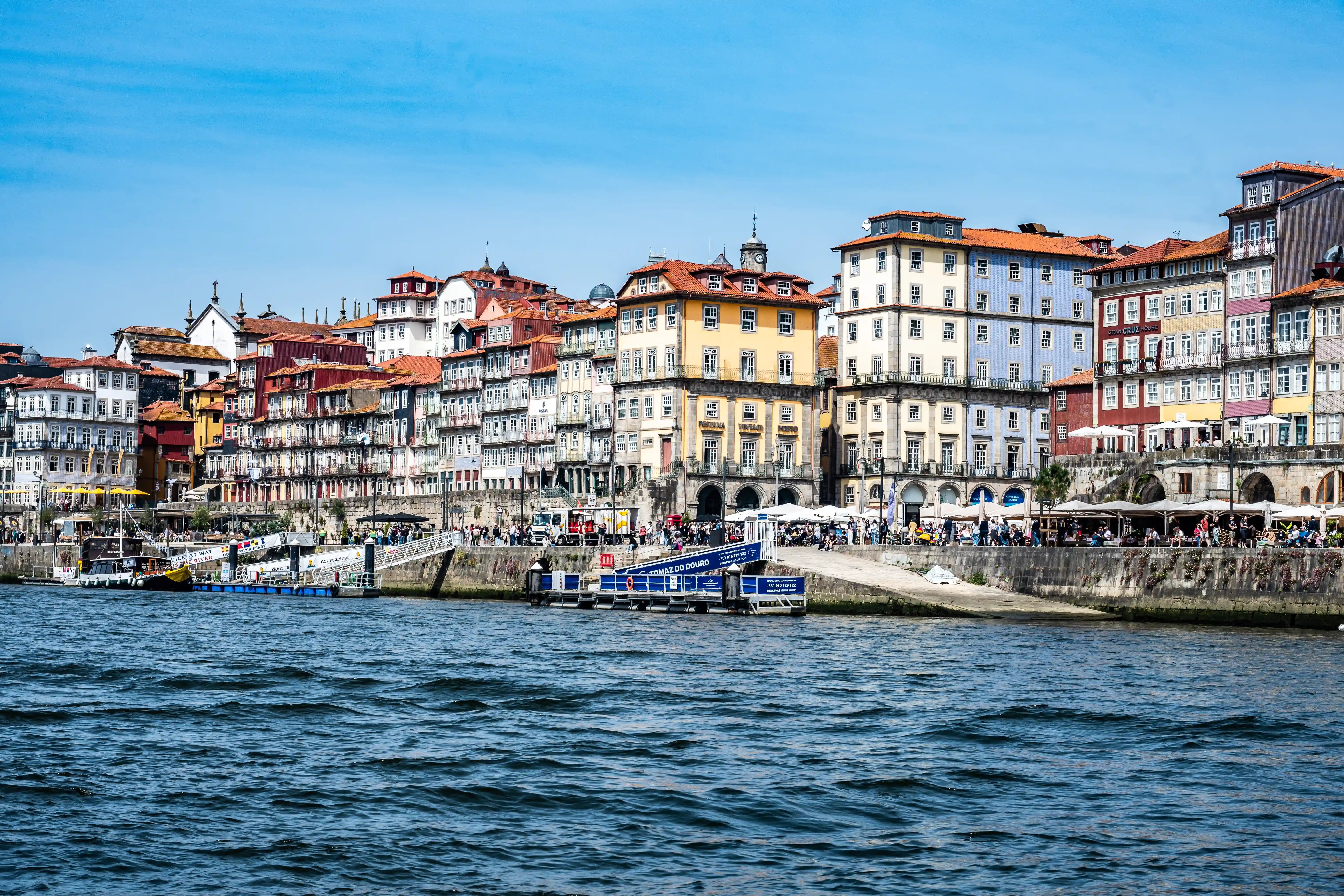 A panoramic view of the Ribeira district in Porto, Portugal, featuring a diverse array of colourful buildings with terracotta roofs facing the blue waters of the Douro River. A bustling promenade with people, cafes under umbrellas, and a blue boat tour pontoon are visible.