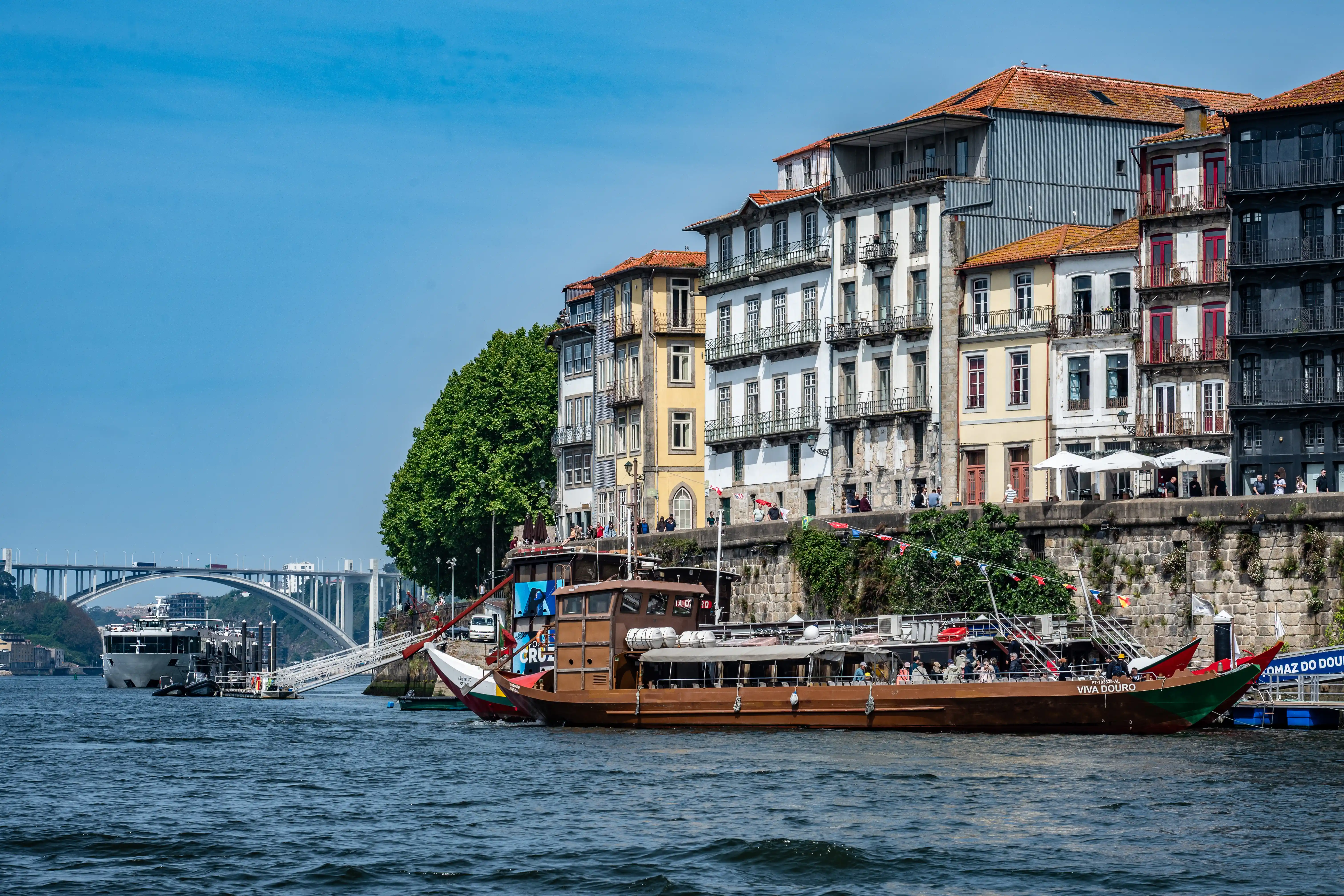 A scenic view of the Douro River in Porto, Portugal, featuring historic buildings with colourful facades and intricate balconies, alongside two boats on the water and a large arch bridge in the background under a clear blue sky.