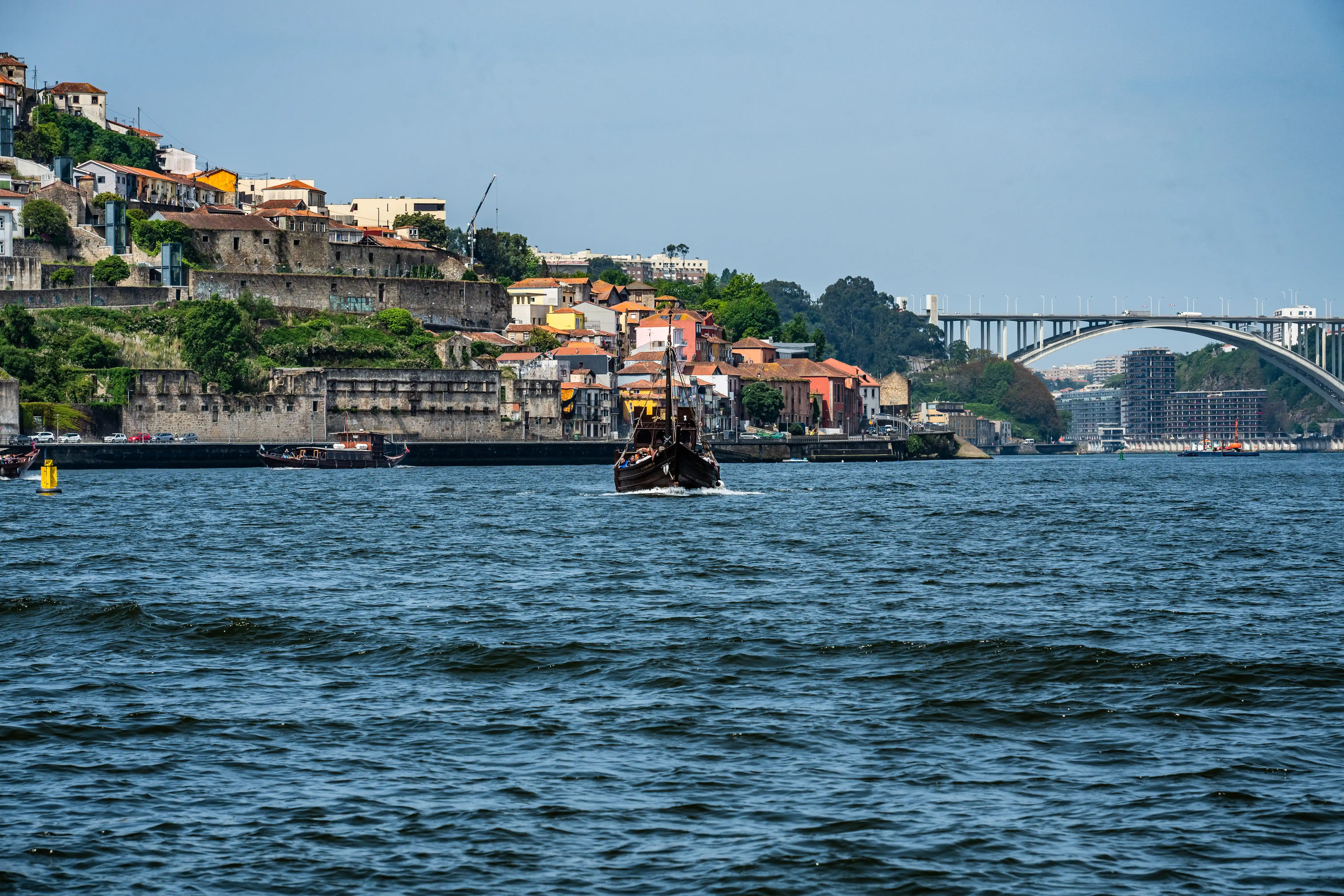 A traditional wooden boat with a prominent sail structure navigates the blue, rippled waters of the Douro River. In the background, the colourful, historic buildings of Porto's Ribeira district rise up the hillside, with the Arrábida Bridge visible in the far distance.