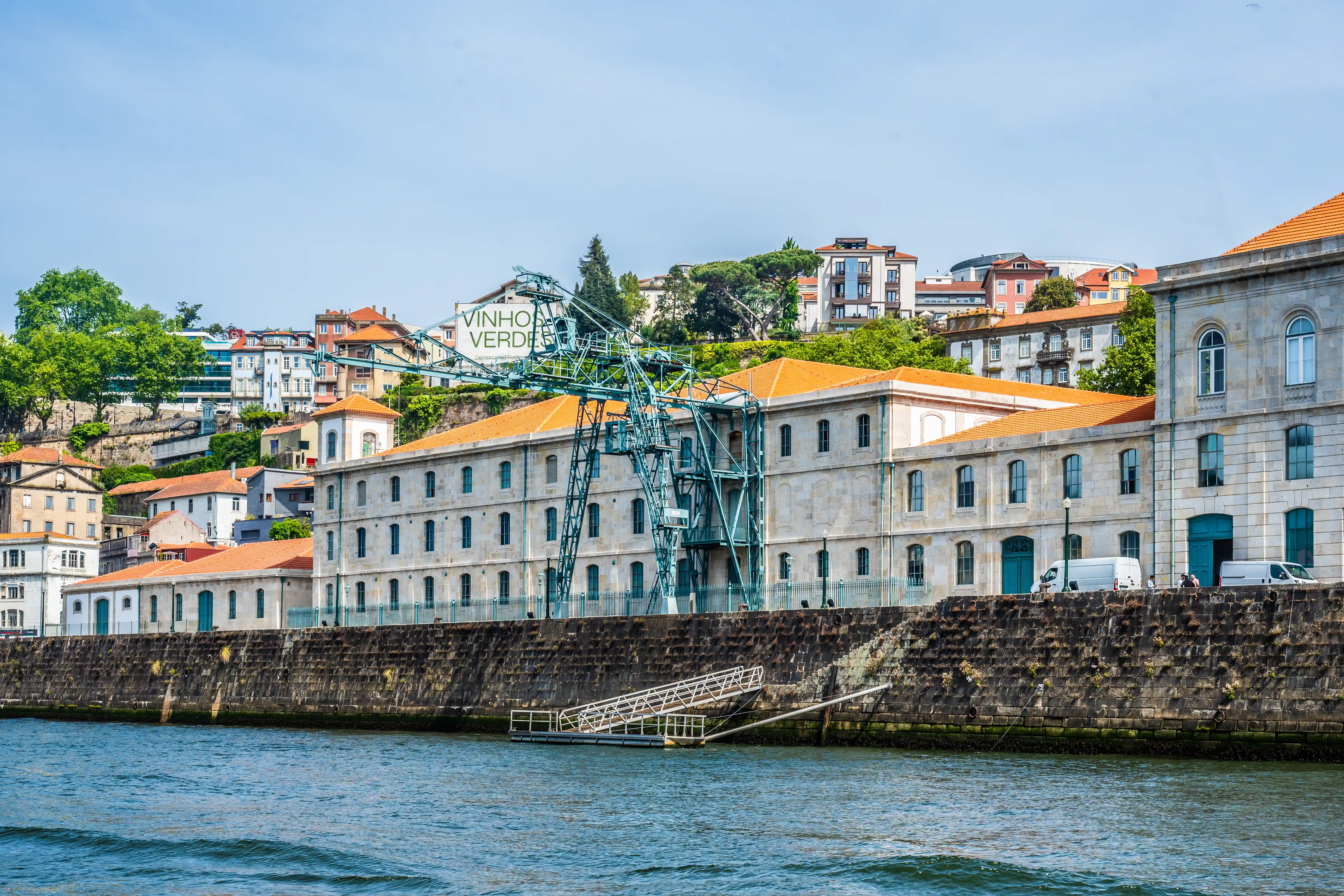 A wide shot of the Douro River with a stone quay in the foreground. Behind the quay are several low-rise, light-coloured stone buildings with orange tiled roofs. A large, teal-coloured industrial crane stands prominently in front of some of these buildings. Further up the hill behind the buildings are more houses and trees under a clear blue sky.