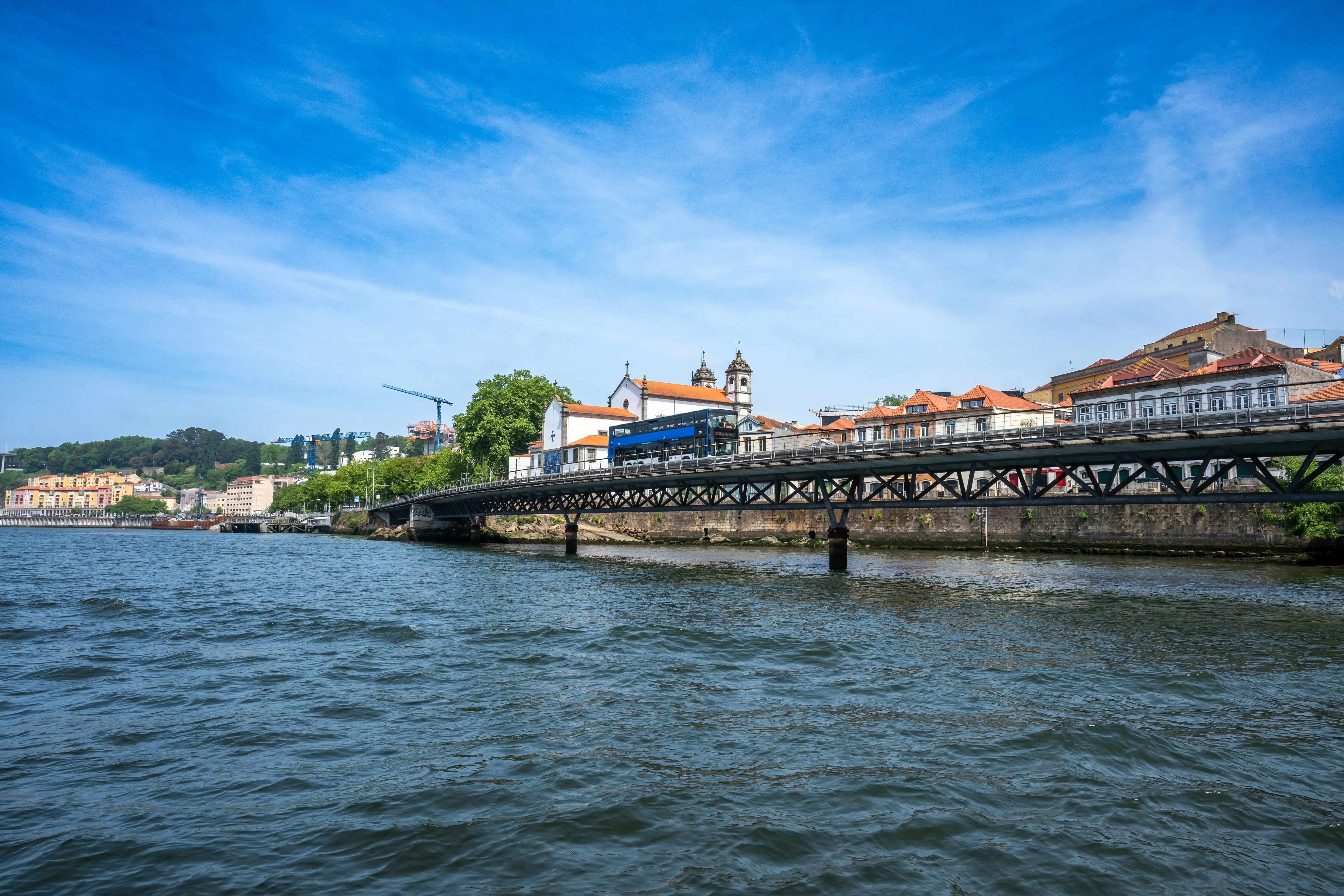 A view from a boat on the Douro River looking towards a metal truss bridge with a blue double-decker bus crossing it. In the background, traditional Portuguese buildings with red tile roofs are visible, along with some trees and construction cranes on a hill under a clear blue sky.