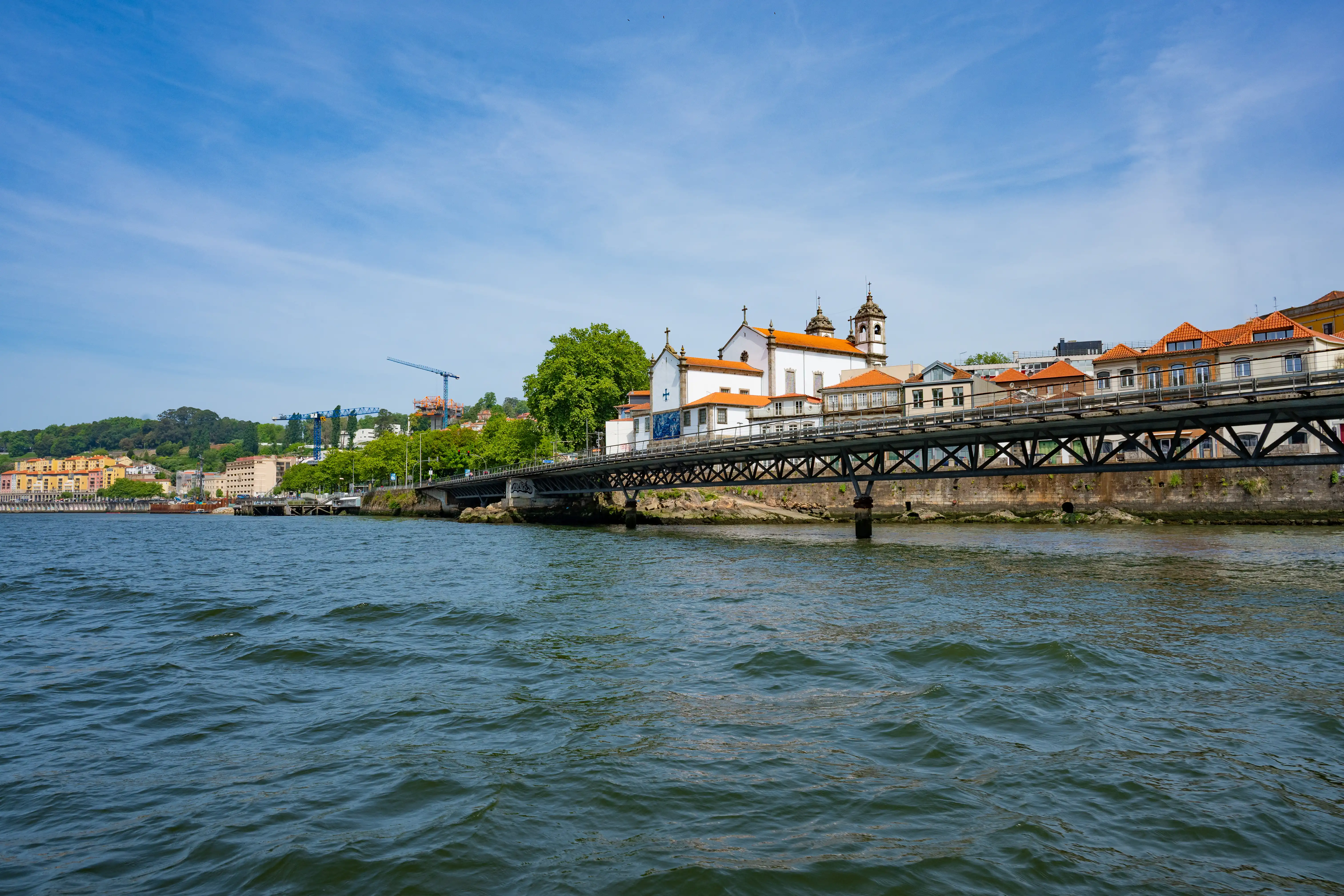 A view across the Douro River in Porto, Portugal, showing the metal structure of the Ponte Dom Luís I bridge, with a white church and other buildings with terracotta roofs lining the riverbank under a blue sky.