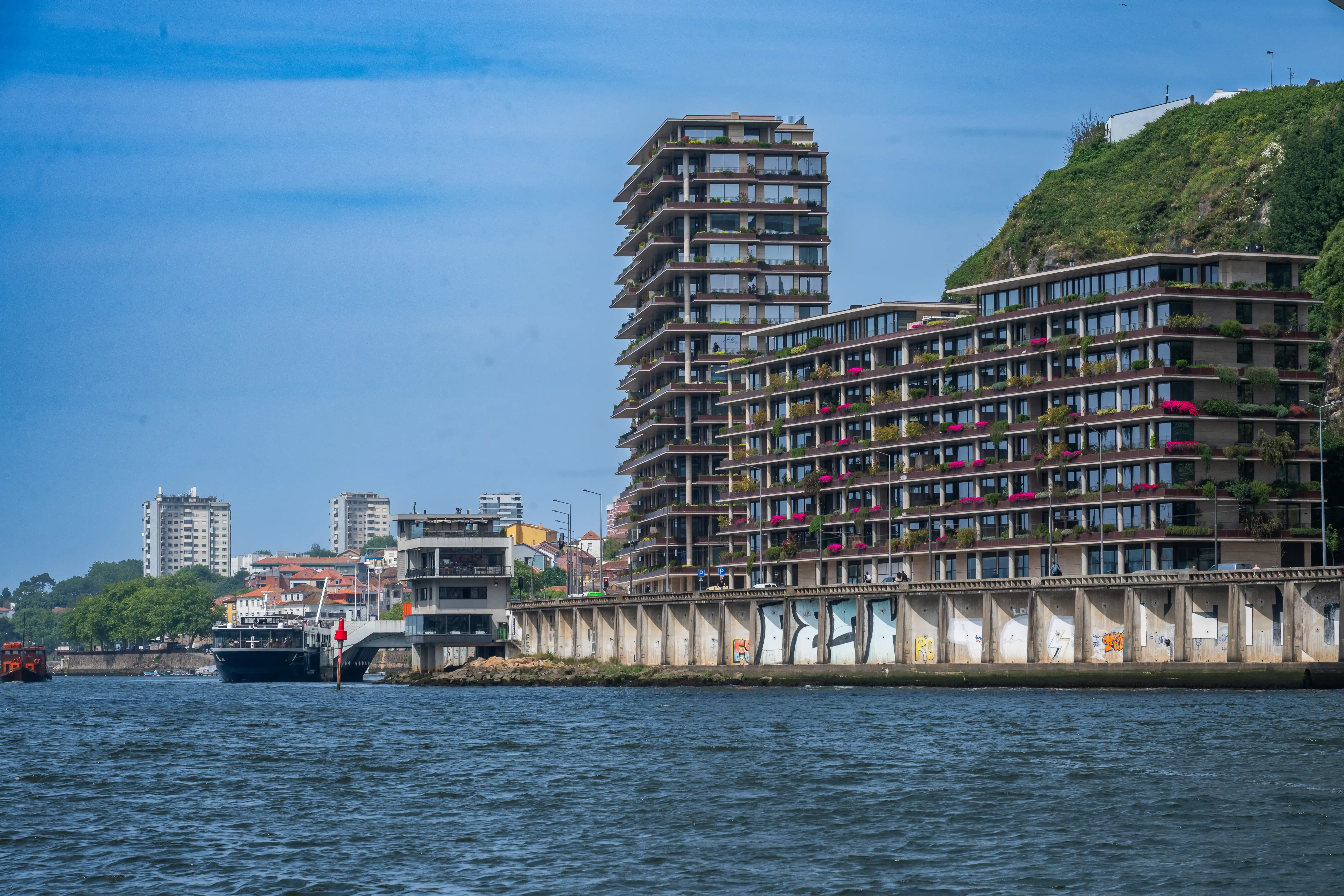A wide shot of a river with modern, multi-tiered apartment buildings featuring many balconies with plants and flowers. A tall, stepped building stands to the left of the lower, terraced buildings. A concrete wall with graffiti separates the buildings from the river. A boat is docked on the left side, and more traditional buildings and a green hill are visible in the background. The sky is a clear blue.