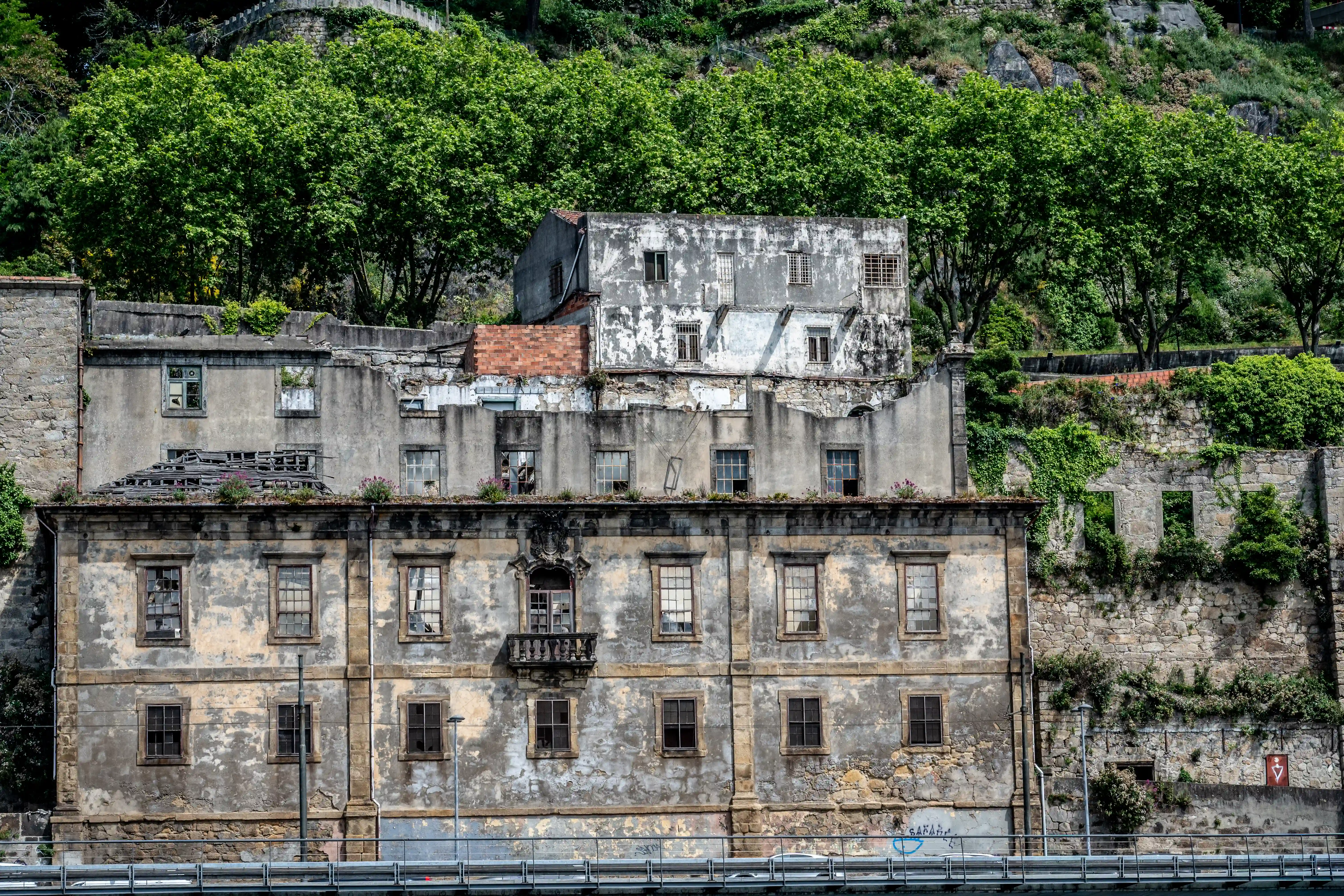 A row of crumbling, abandoned buildings with broken windows and peeling plaster, set against a backdrop of dense green trees and overgrown stone walls.