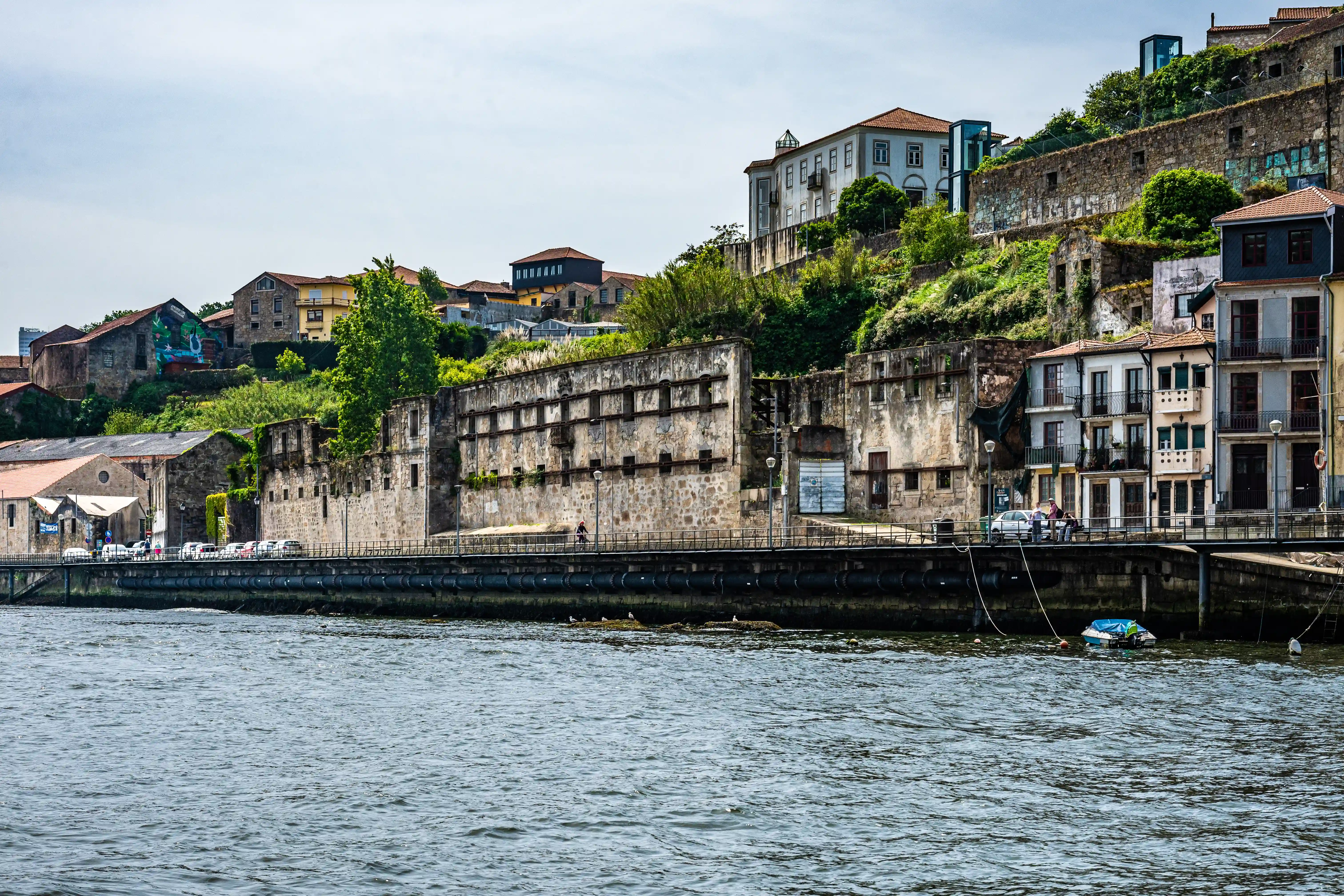 A wide shot of a river with a stone embankment and a row of old, textured buildings on the left, some with weathered facades. Further up a steep hill, more colourful houses with red-tiled roofs are nestled amongst lush green trees and vegetation. A metal railing runs along the embankment, with a few people and cars on the path above.