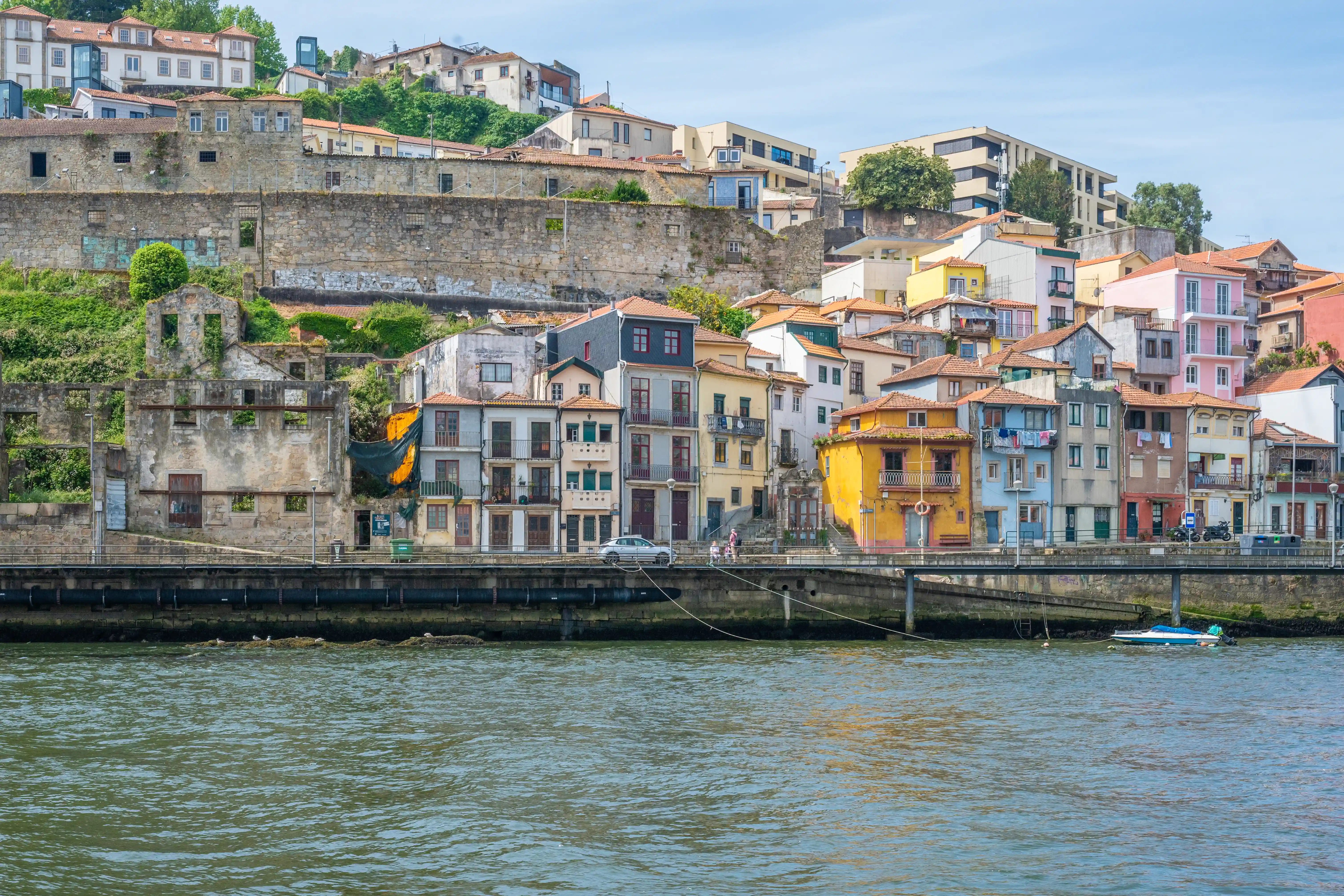 A panoramic view of the colourful buildings of Porto's Ribeira district, lining the bank of the Douro River, with a bridge, a car, and a small boat.