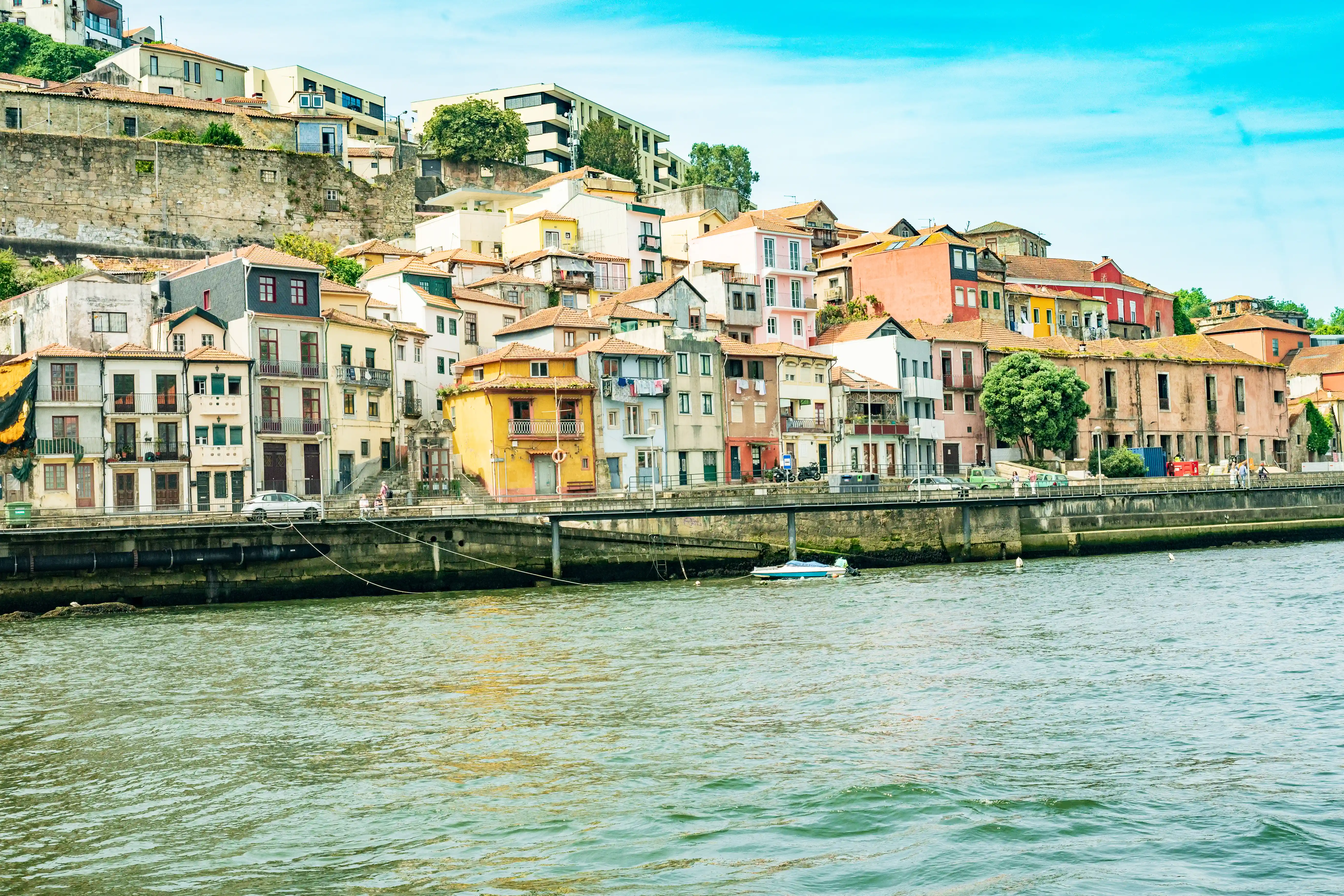 A panoramic view of the colourful buildings of Porto's Ribeira district, lining the bank of the Douro River, with a bridge, a car, and a small boat.