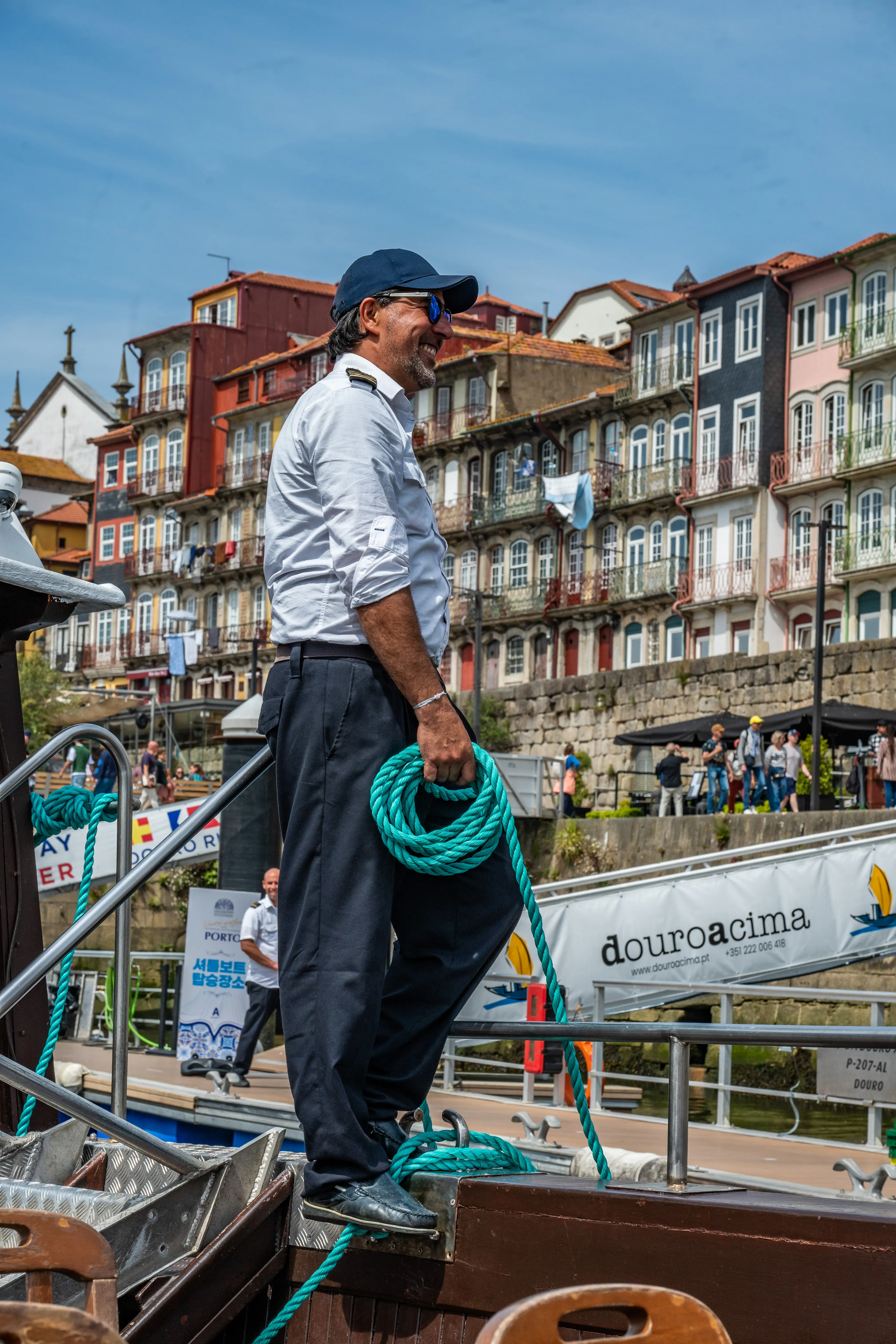 A smiling man in a captain's attire, holding a coiled turquoise rope, stands on a boat with a backdrop of colourful buildings along a river.