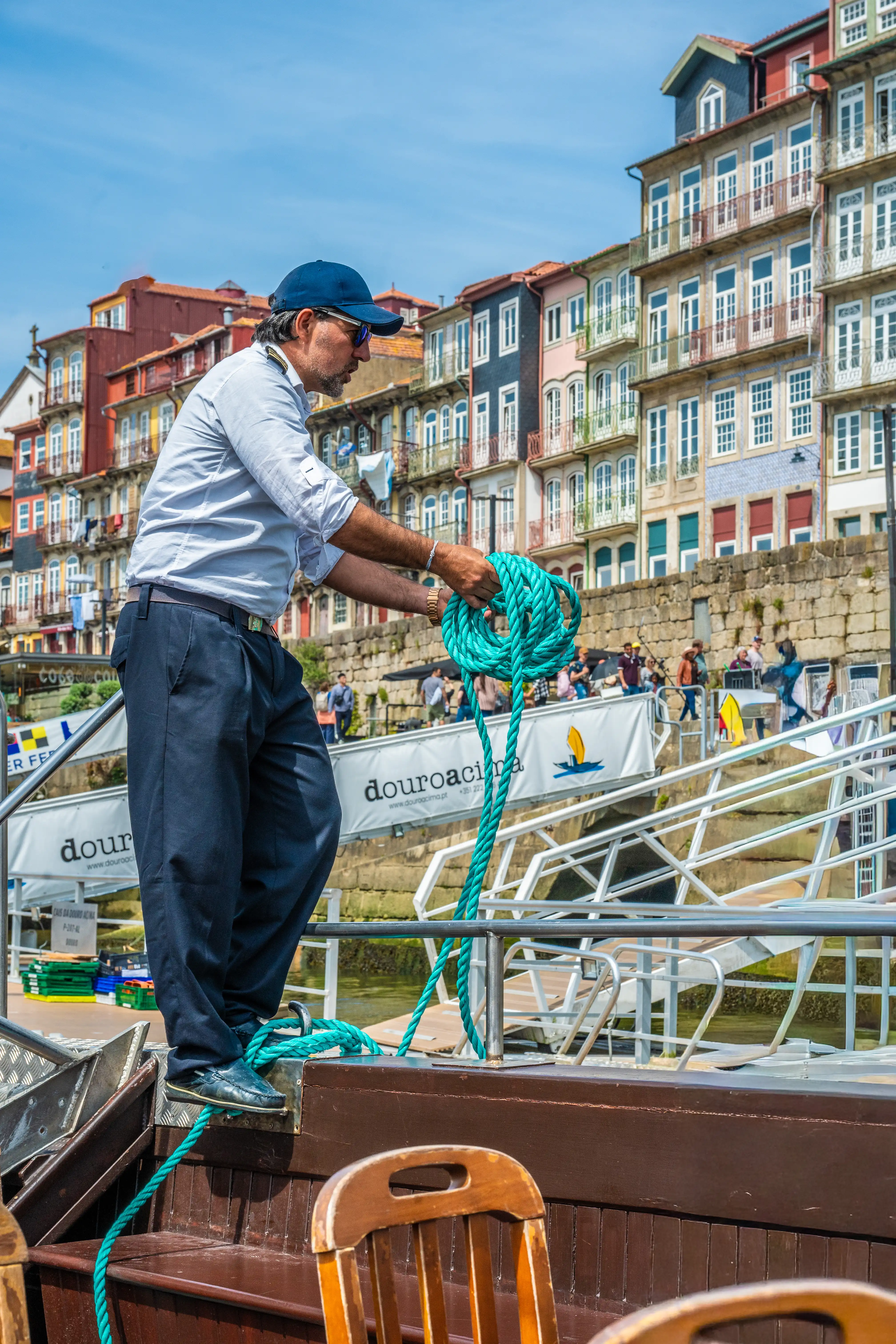  A man in a blue cap and light shirt coils a thick turquoise rope on a boat deck, with the colourful buildings of Porto's riverfront and a blue sky in the background.