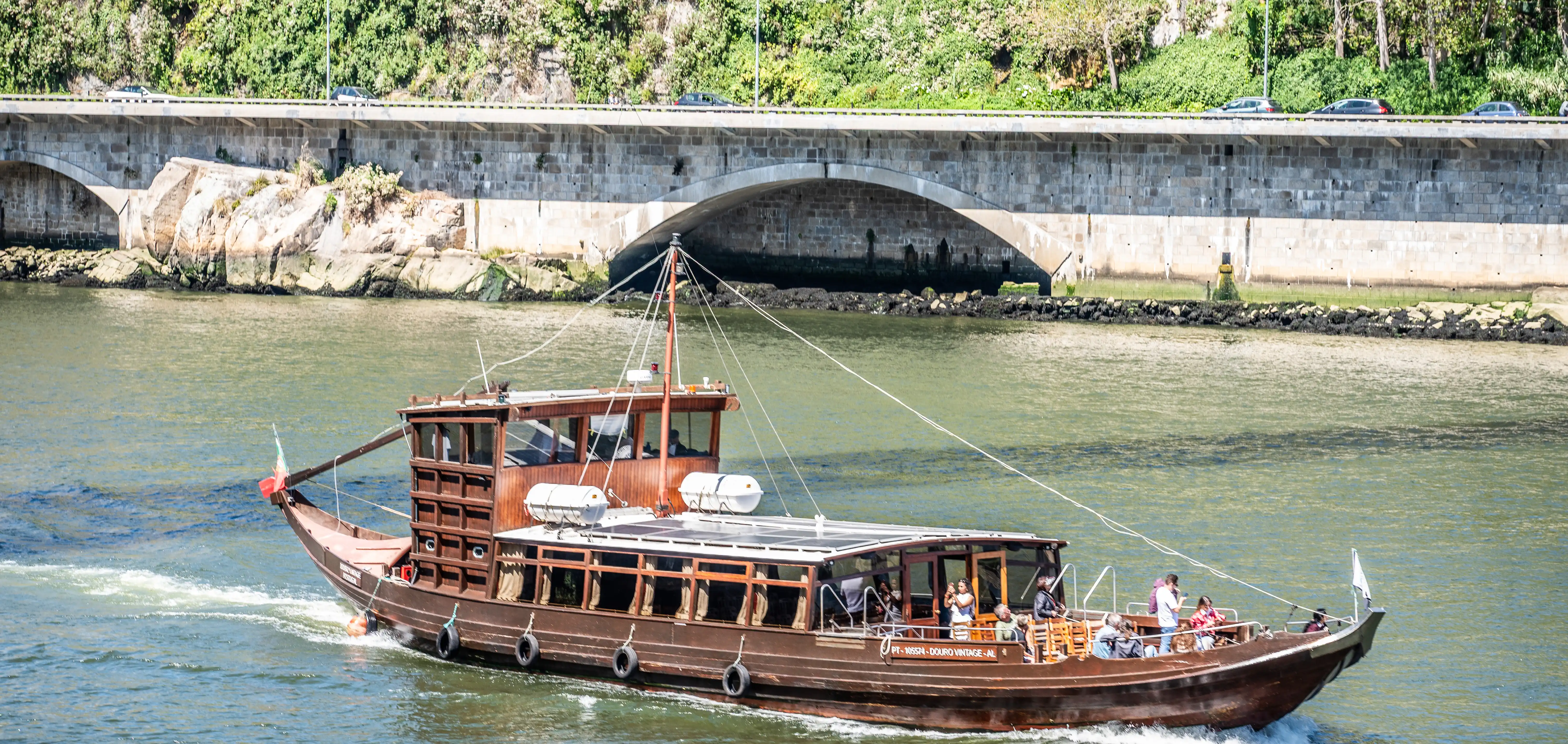 A brown wooden tour boat with solar panels on its roof sails on the Douro River, passing under a large stone arched bridge. People are visible on the deck of the boat, and cars drive on a road above the bridge.