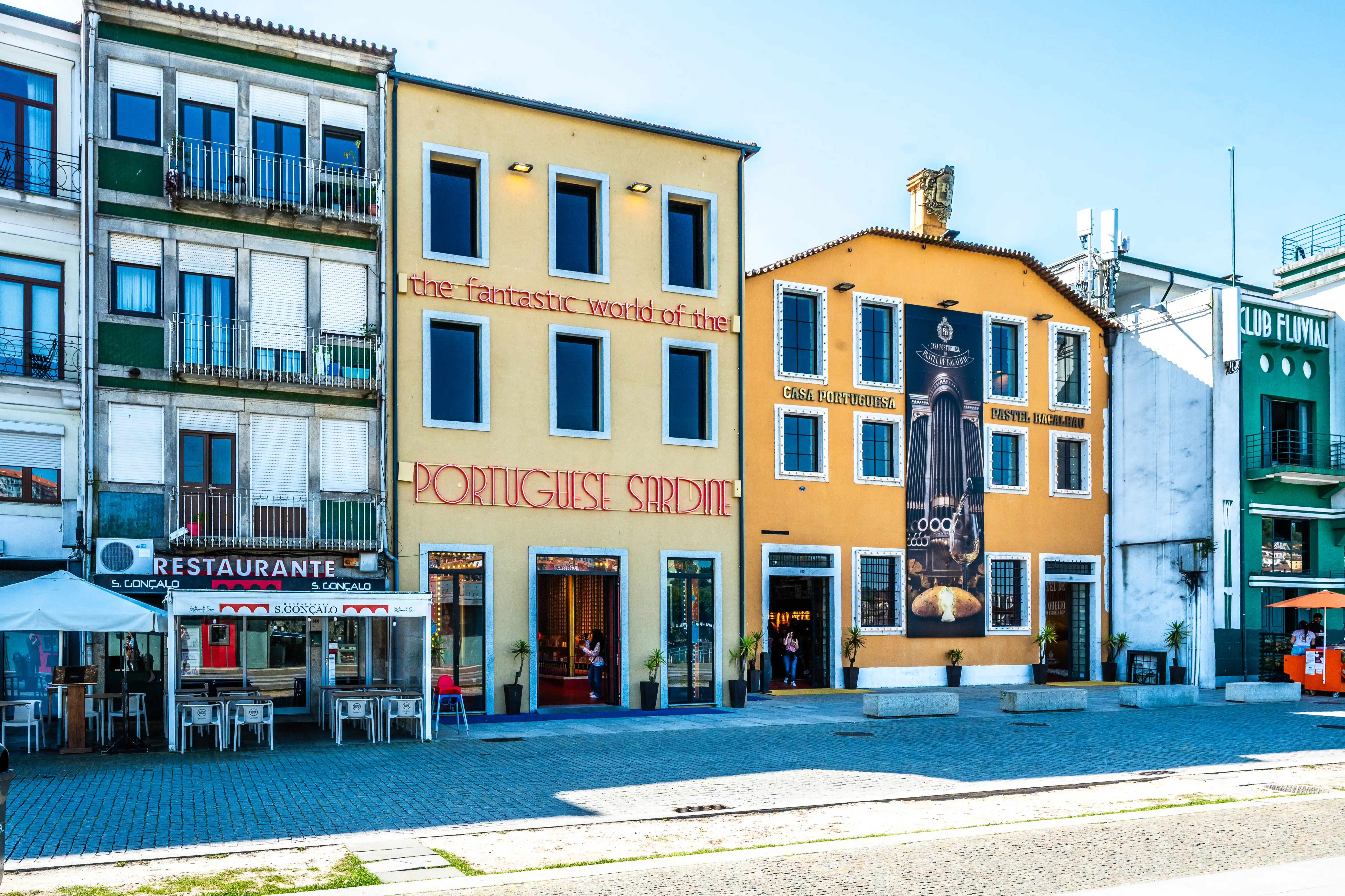 The image depicts a vibrant street scene featuring a corner building with multiple businesses. The most prominent sign reads 'PORTUGUESE SARDINE,' indicating a seafood restaurant. Adjacent businesses include a restaurant named 'S. Gonçalo' and a 'Pastelaria,' suggesting a lively area with dining options. The architecture is characterized by colorful, multi-story buildings with balconies and storefronts, contributing to a bustling urban atmosphere