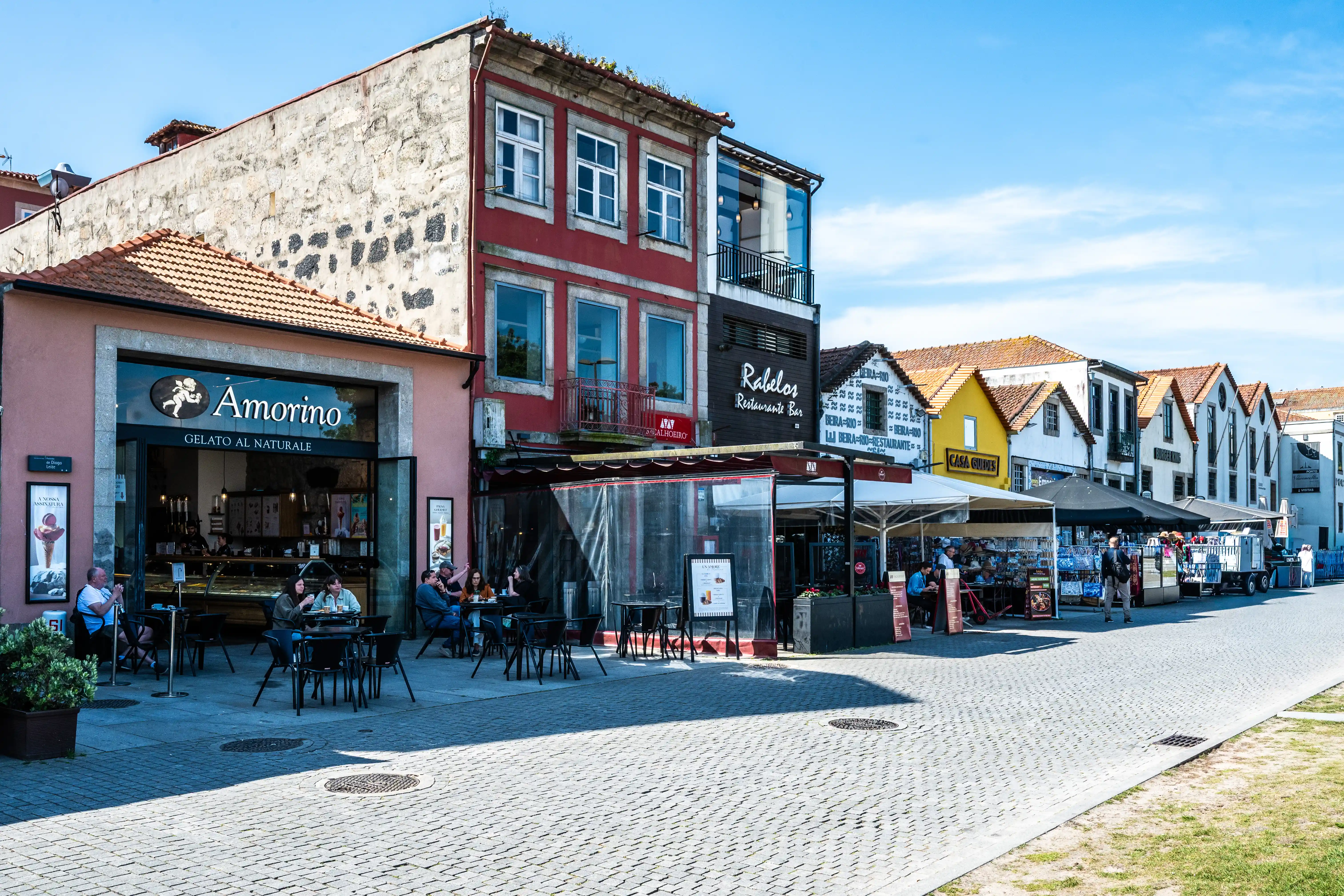 he image depicts a quaint street scene in what appears to be a European town, featuring a gelato shop named 'Amorino' with outdoor seating for patrons. Nearby, there's a restaurant called 'Rabelo Restaurante Bar.' The street is lined with charming, traditional buildings, and a few pedestrians are visible, enjoying the pleasant weather under a clear blue sky.