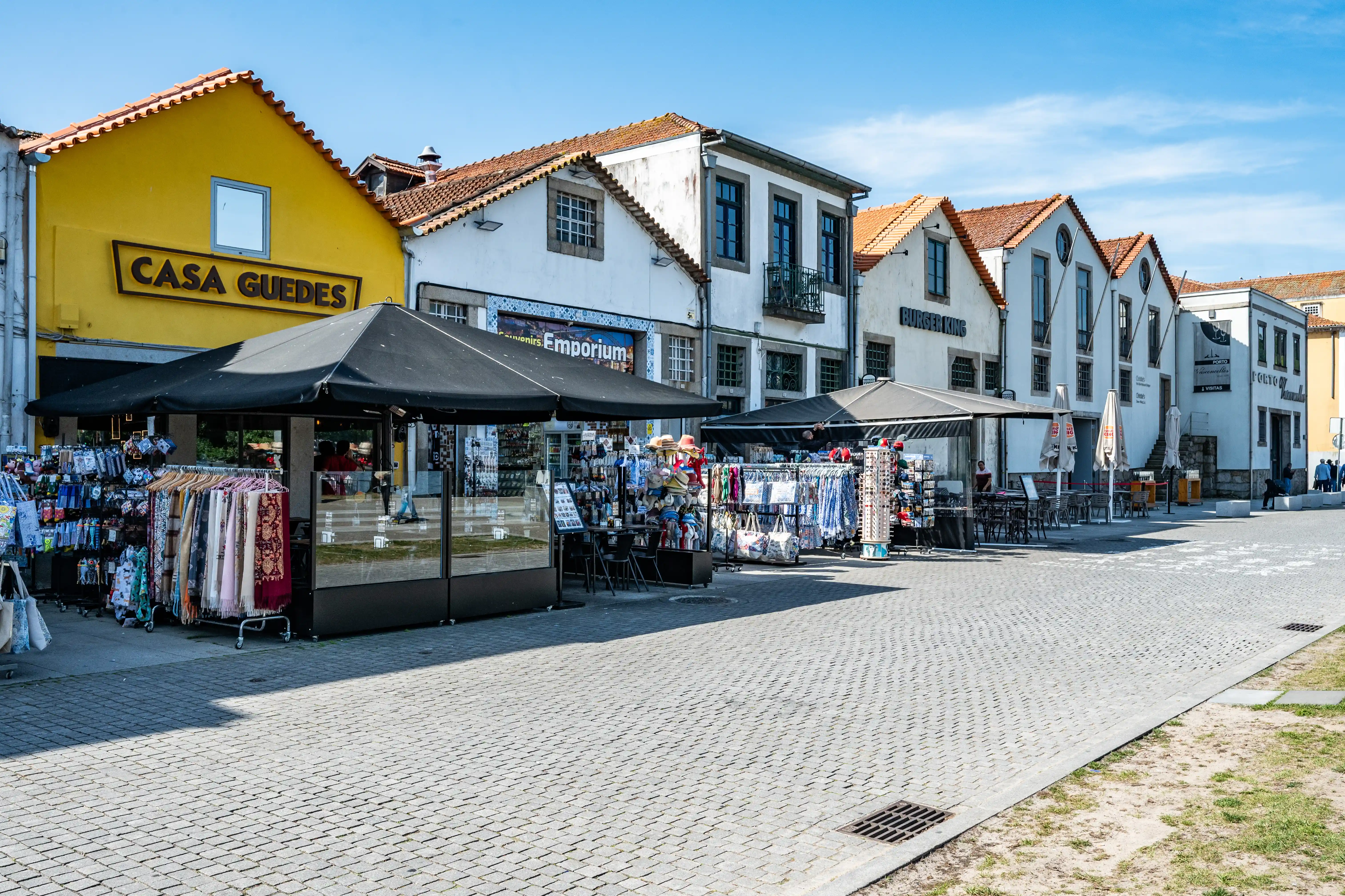 The image depicts a quaint European street scene with a row of charming, pastel-colored buildings housing various shops and boutiques. The establishment 'Casa Guedes' is prominently visible, featuring a black awning and a yellow facade. The street is paved with cobblestones, and a few pedestrians can be seen strolling along the sidewalk. The overall atmosphere is lively and inviting, suggesting a bustling commercial area.