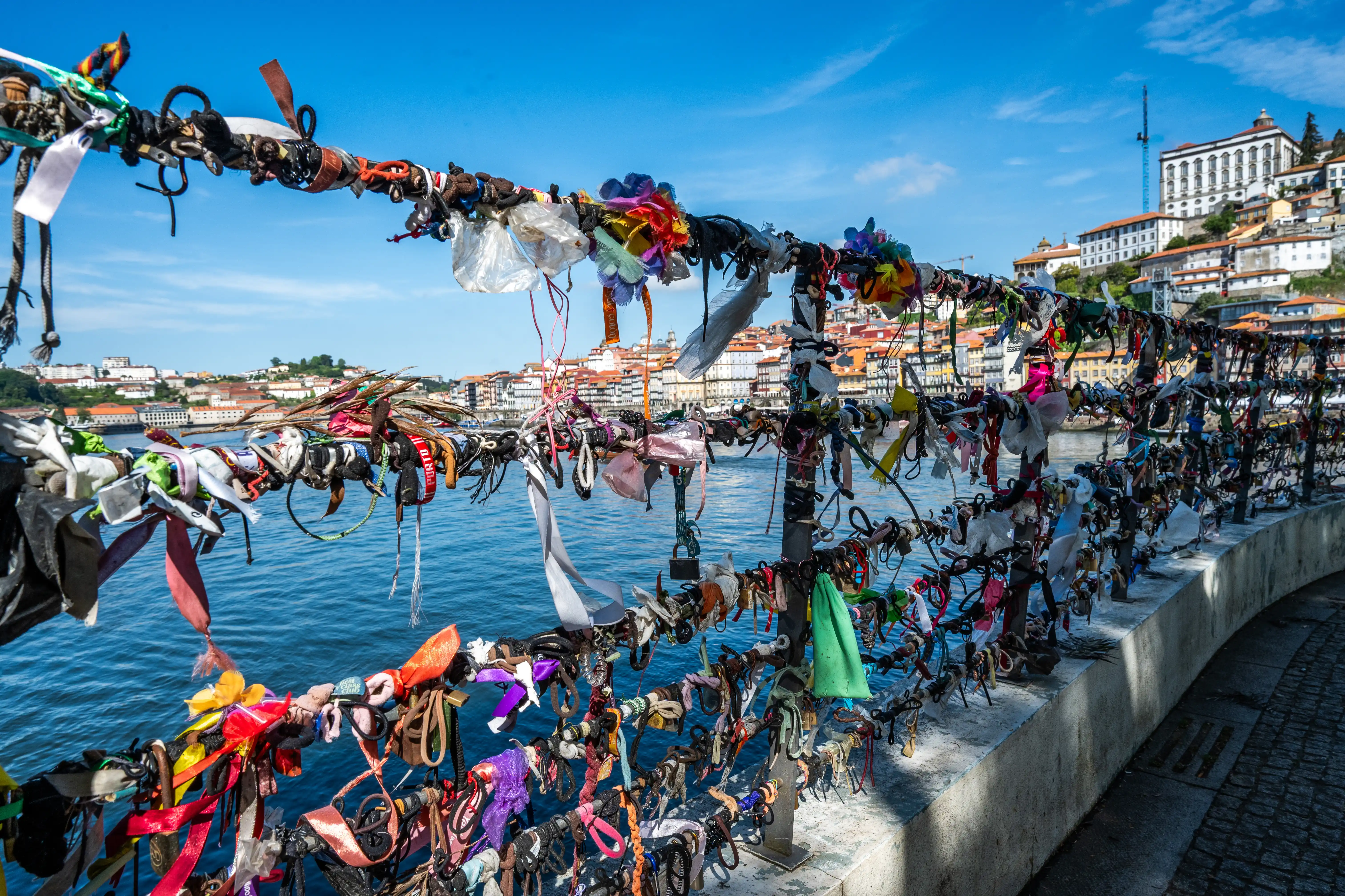 A close-up view of a railing covered in numerous colourful love locks and ribbons, with the city of Porto and the Douro River in the background..