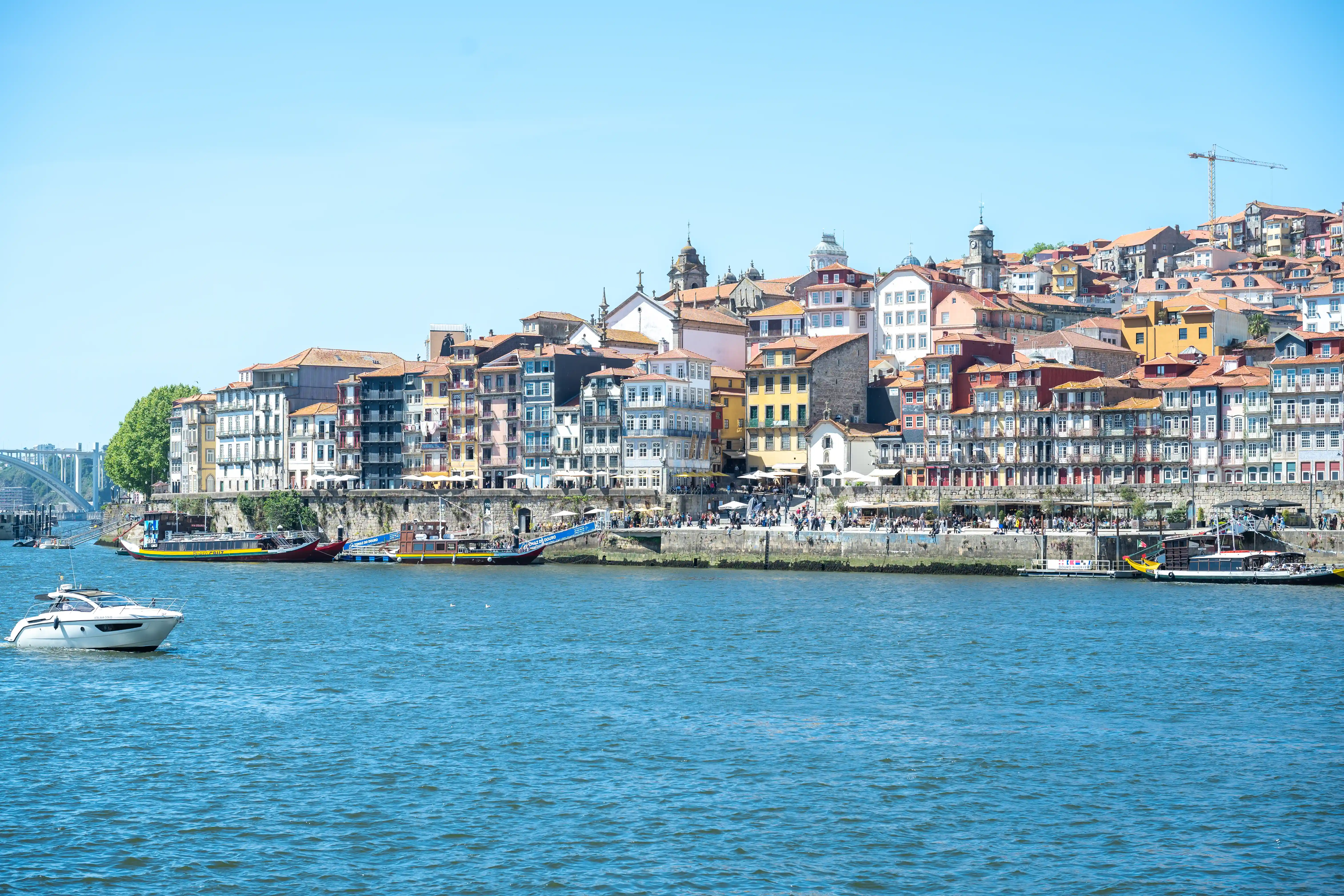 The image depicts a vibrant coastal cityscape featuring a bustling waterfront lined with colorful buildings. The buildings, varying in architectural styles and colors, extend up a hillside, creating a picturesque backdrop. Several boats and yachts are docked along the waterfront promenade, which is crowded with people. The clear blue water adds to the scenic beauty, and a small motorboat is cruising in the foreground.