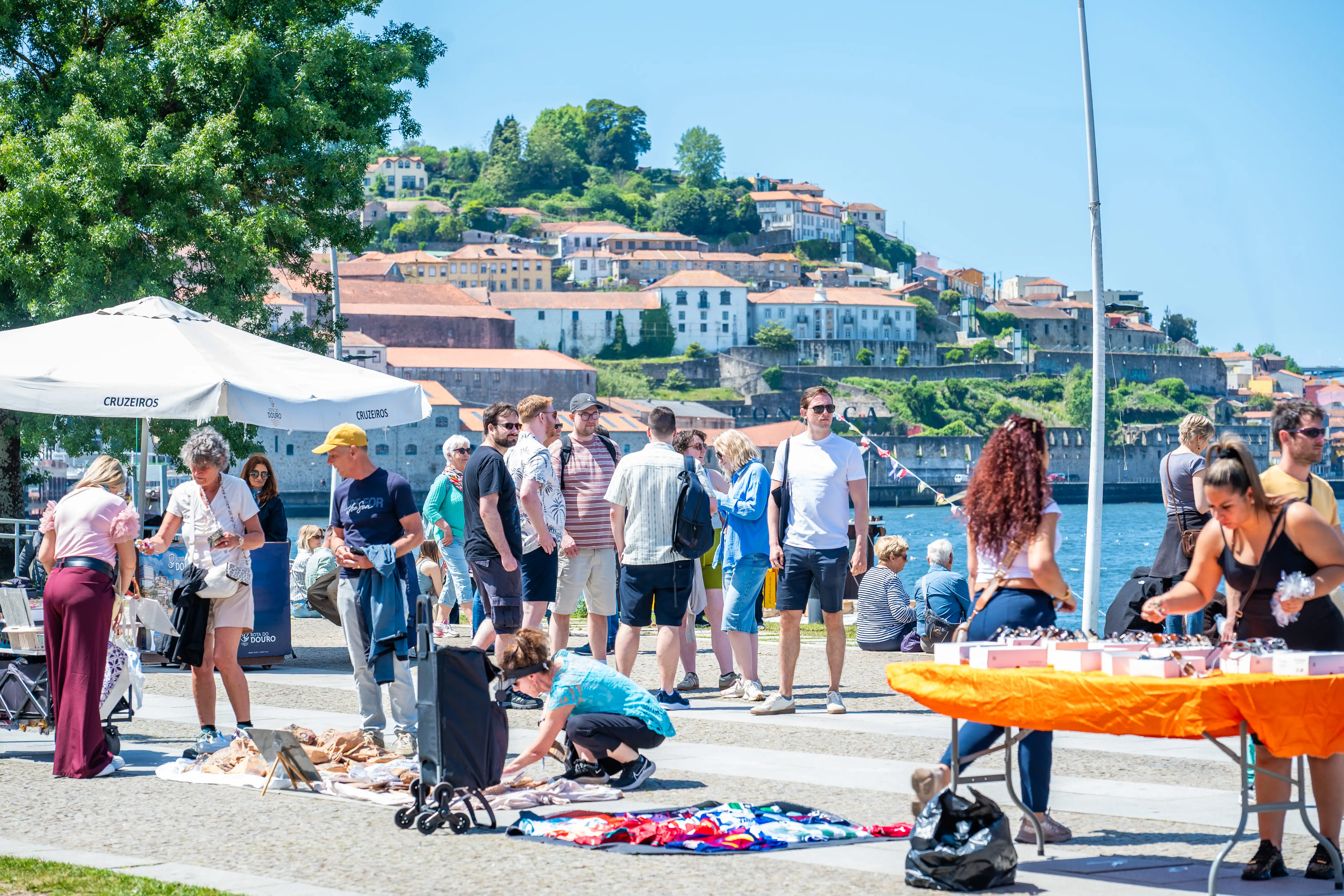 The image depicts a lively outdoor scene where a group of people gathers around a large inflatable slide. The setting appears to be a sunny day, with a scenic backdrop of a coastal town featuring white buildings and lush greenery. The crowd includes adults and children, many wearing casual summer attire. Some people are interacting with the slide, while others are in conversation or observing. The atmosphere suggests a community event or festival.