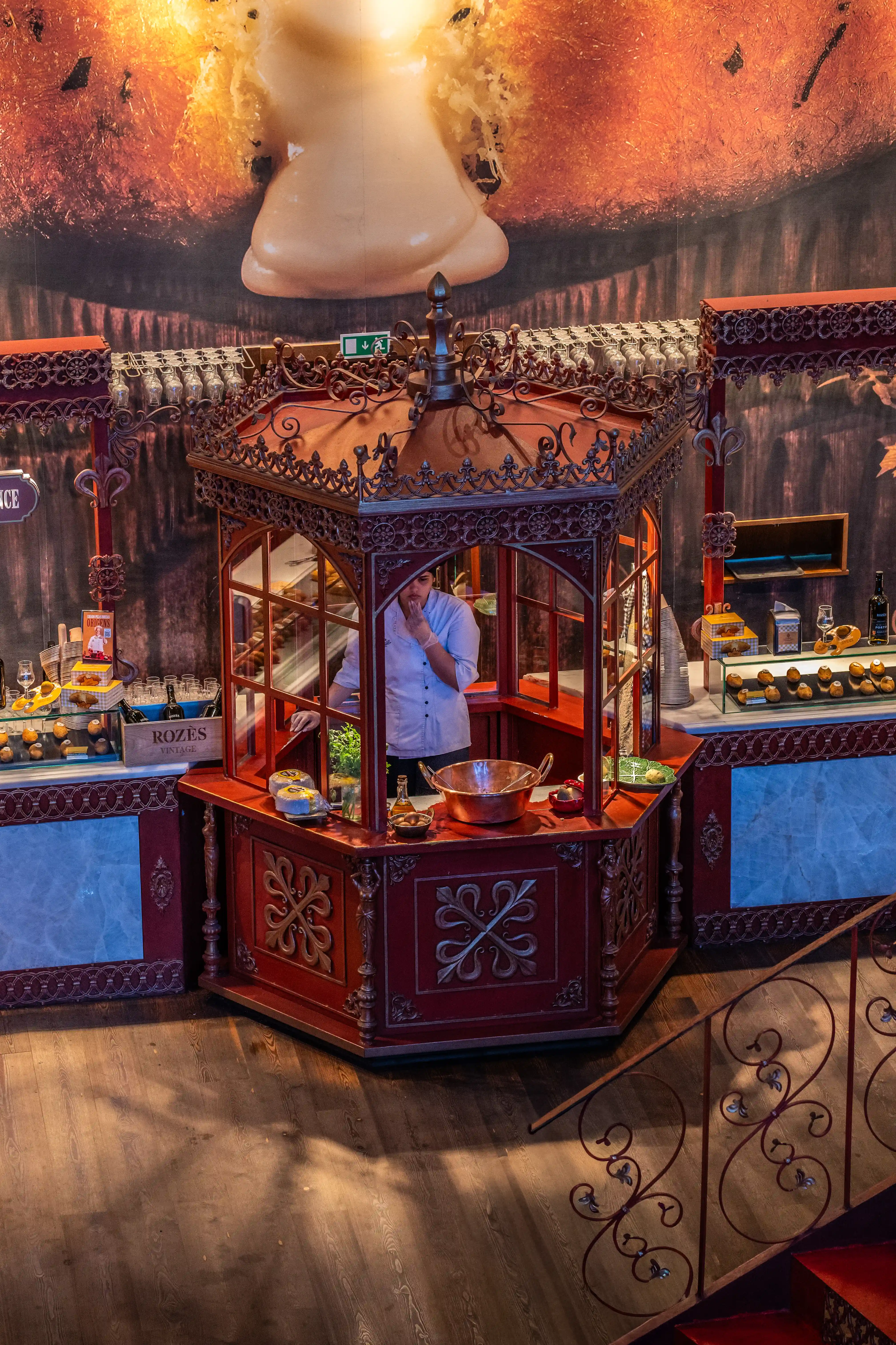 Ornate red and gold food stall with a chef inside preparing pastries, displayed with various baked goods, under a large abstract mural with a staircase on the side.