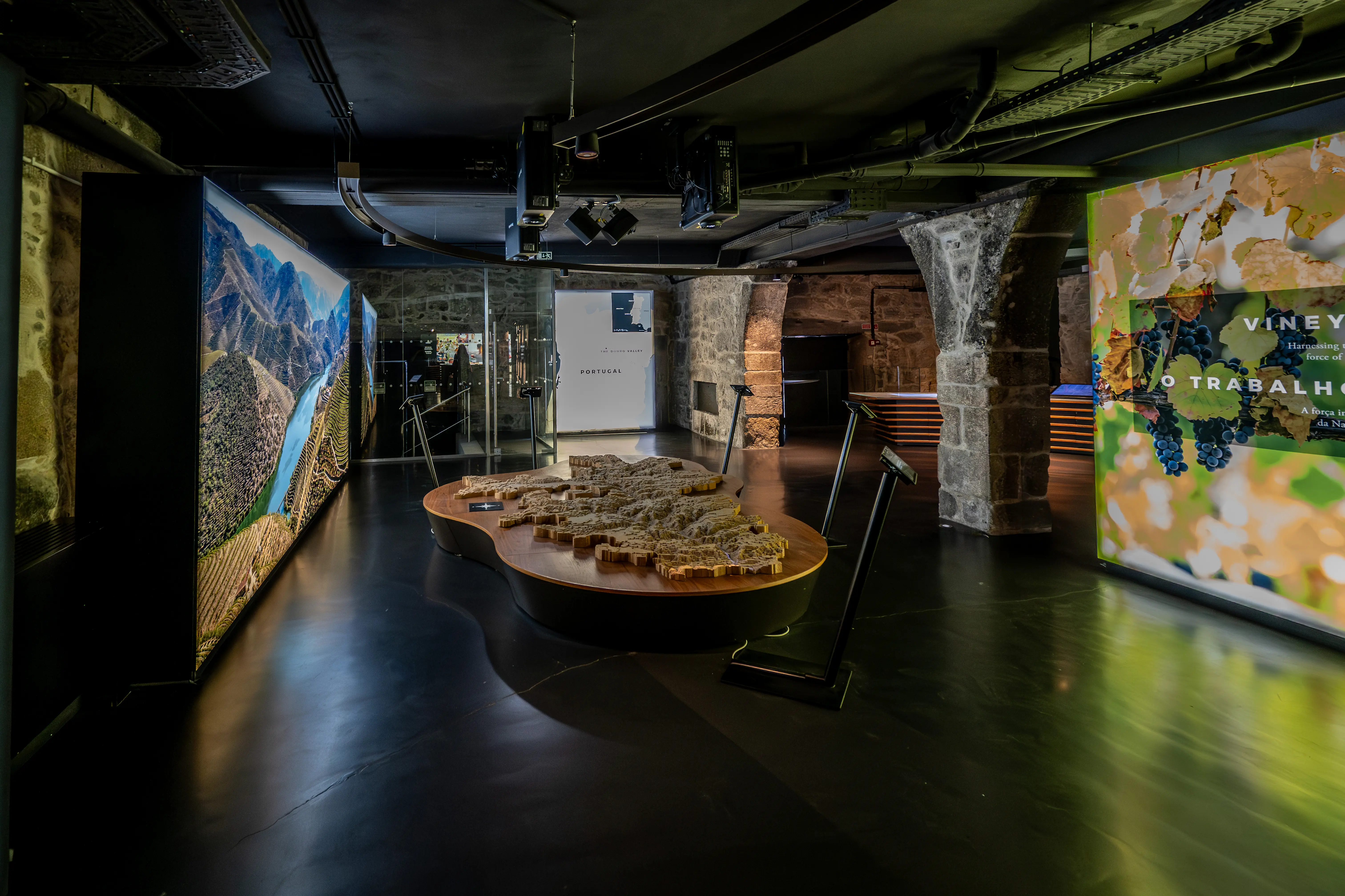 The image depicts an indoor exhibit featuring a detailed model of a castle or fortress, likely a historical or cultural landmark. The model is displayed on a raised platform with a circular railing for observation. Surrounding the model are informational posters and images, providing context about the site. The exhibit is well-lit and appears to be part of a larger museum or educational display.