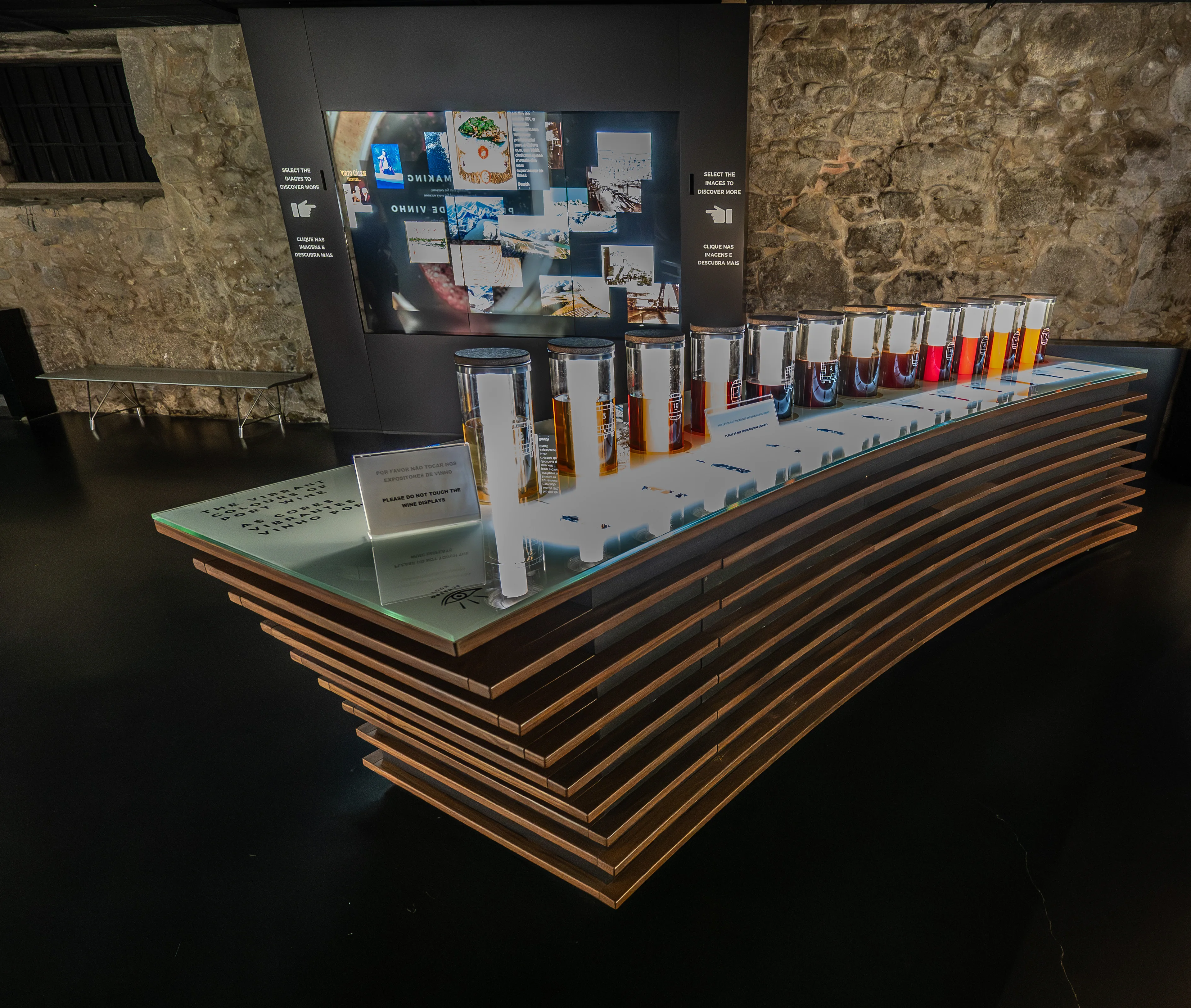 The image depicts an interactive wine tasting exhibit in a museum or educational setting. The display features a touchscreen kiosk with instructions in multiple languages, a wooden counter with multiple wine glasses, and informational panels. A sign in front of the glasses warns visitors not to touch the wine displays. The setup appears designed to engage visitors in learning about wine through sensory experience.