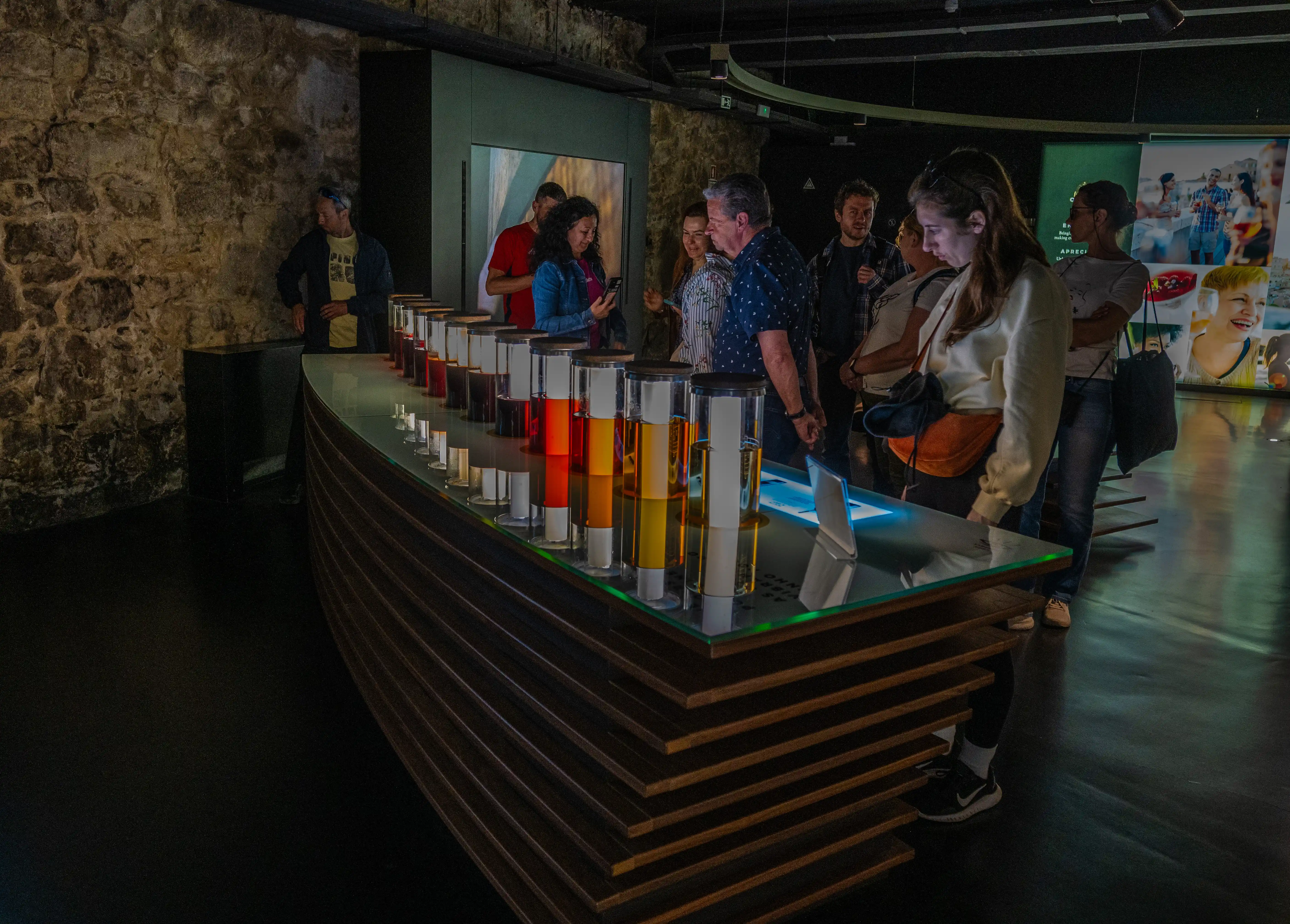 A group of people are gathered around a curved display table in a museum, observing a series of cylindrical glass containers filled with liquids of varying colours, from deep red to vibrant yellow. The display appears to be interactive, with some visitors leaning in to examine the samples and a tablet-like screen visible on the table. The background features stone walls and an exhibit area with large visual displays.
