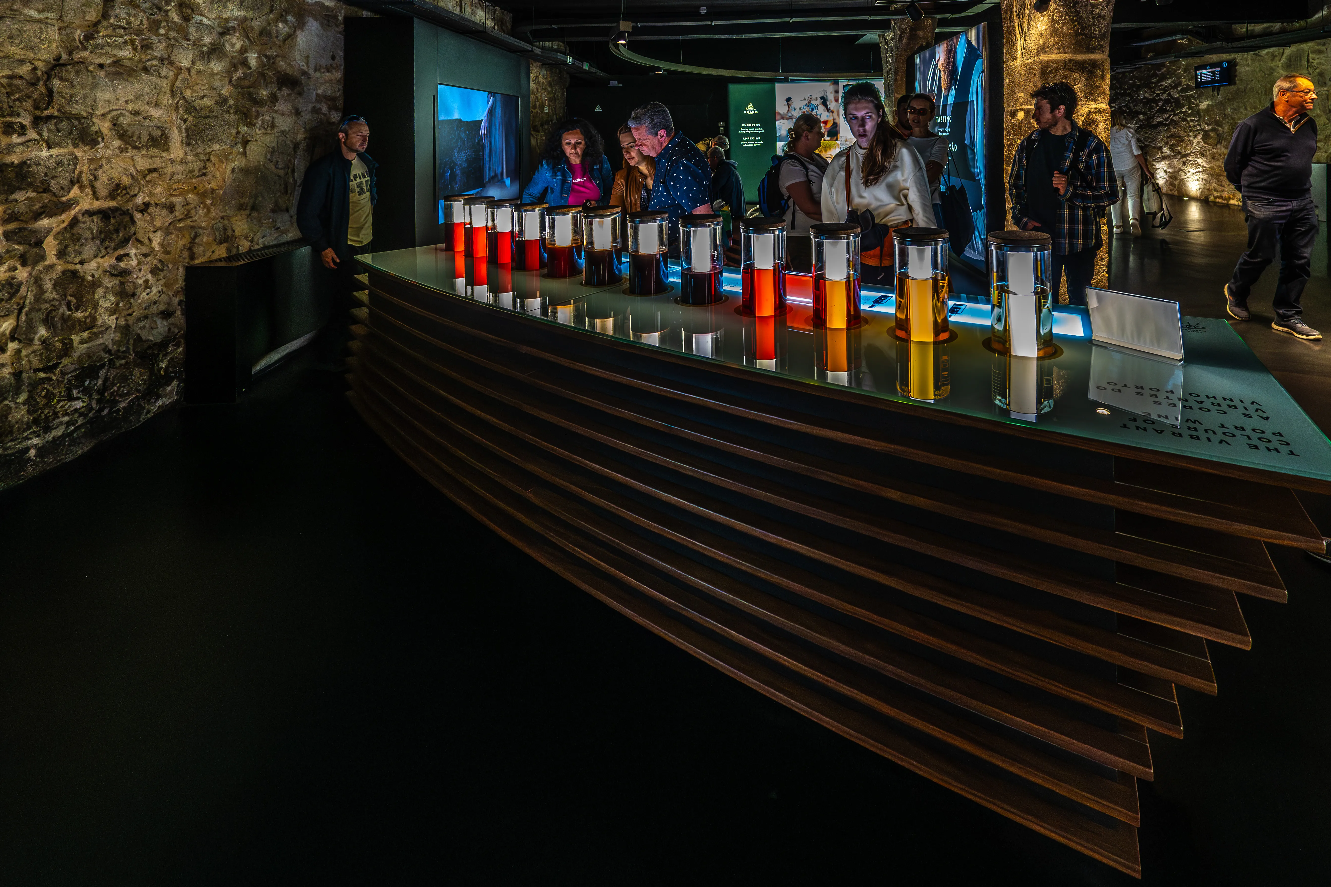 A group of people stand around a display counter featuring multiple glass beakers filled with liquids of varying shades of red, amber, and yellow, representing different Port wine colours. The counter is set against a backdrop of rustic stone walls and modern exhibition elements.