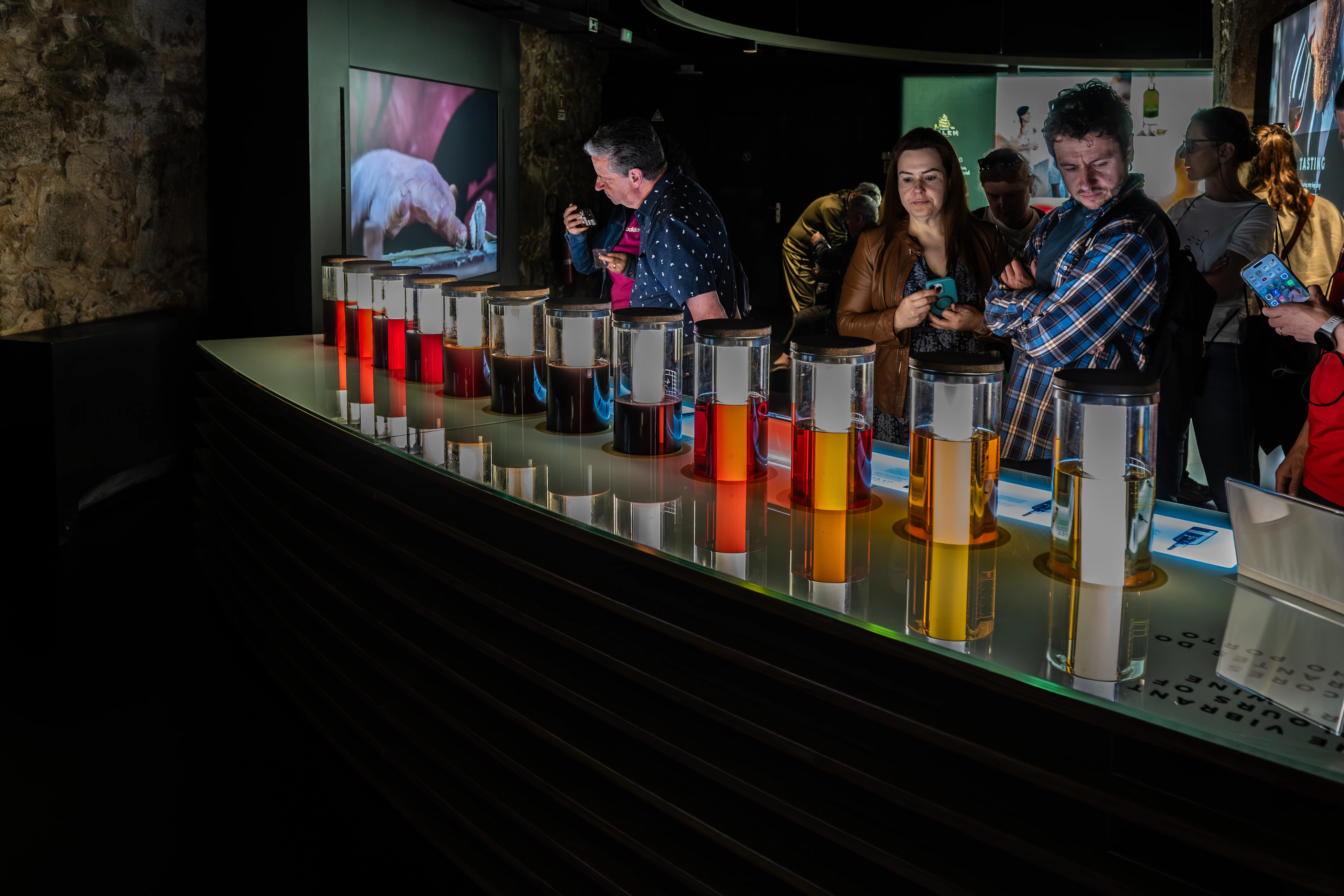 The image depicts a group of people gathered at a bar, socializing and conversing. The bar has a modern design with colorful, illuminated glass shelves and stools. The patrons appear to be enjoying drinks and snacks, with some holding glasses. In the background, a large screen displays an image of a person, suggesting a lively and possibly themed atmosphere in the establishment.