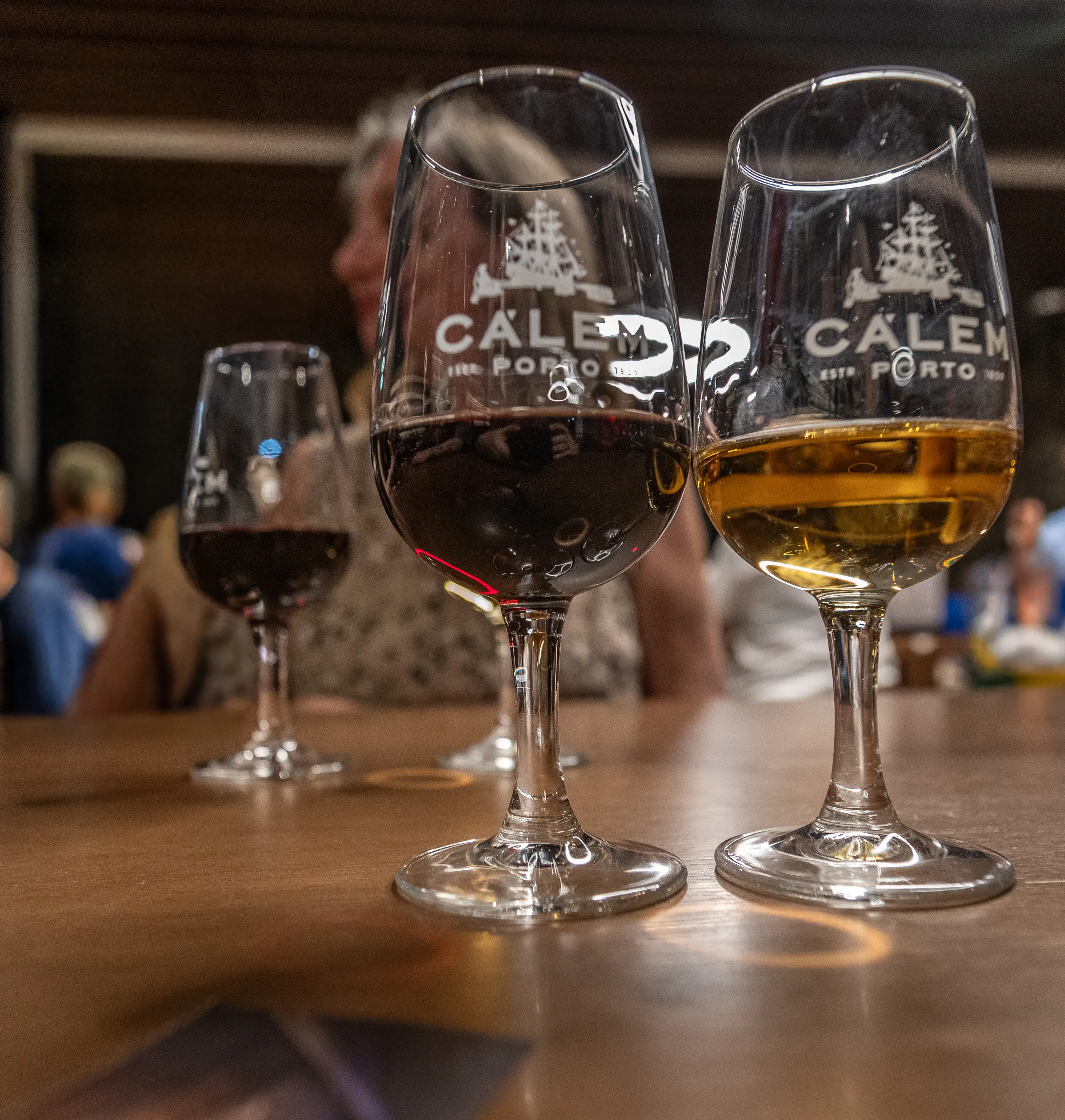 Two elegant wine glasses, branded with the Calem Porto logo, are positioned on a polished wooden surface. The glass on the left contains a deep red port, while the glass on the right holds a golden, amber-coloured white port. The background is softly blurred, showing indistinct figures of people, hinting at a relaxed tasting environment.
